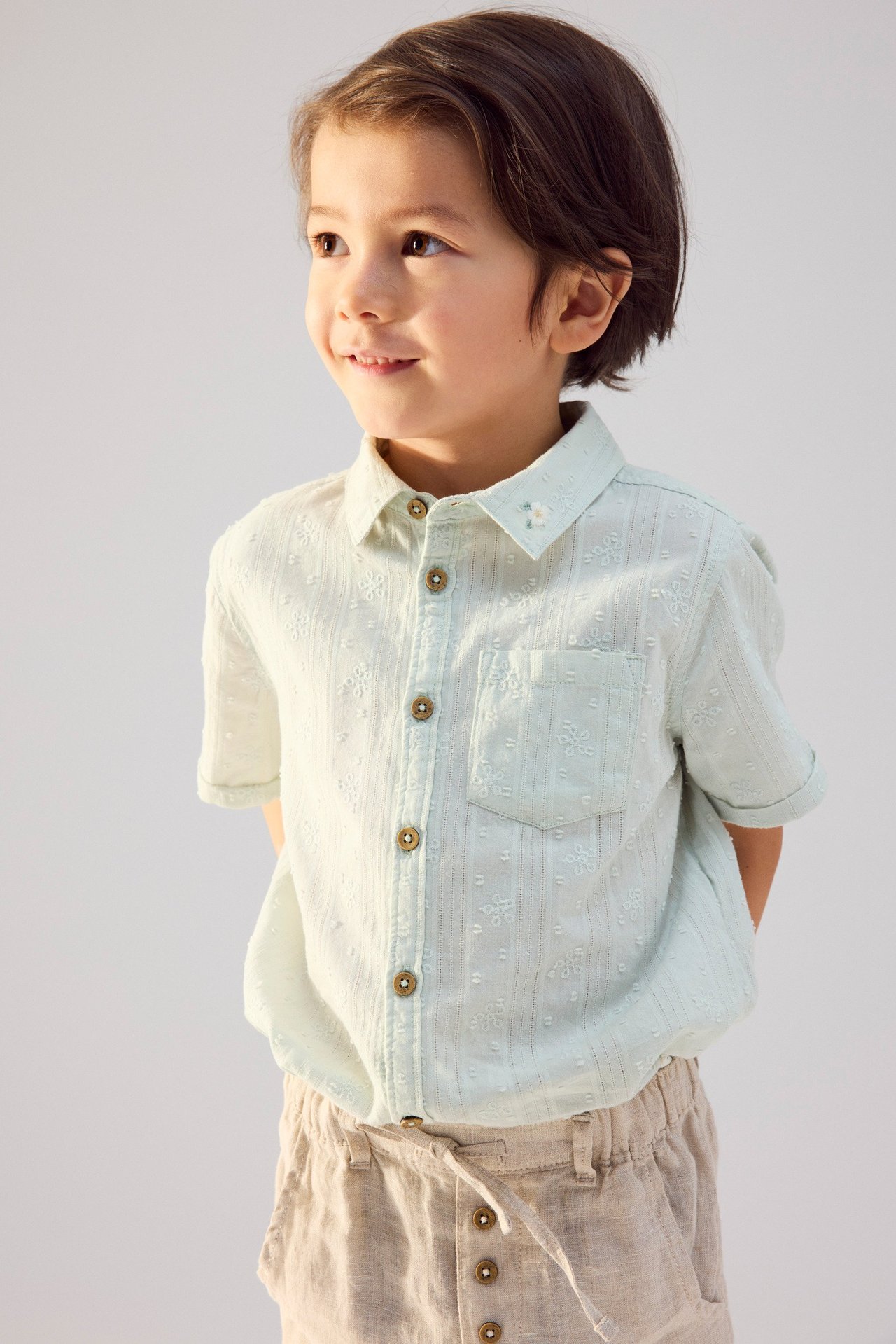 Widok z przodu: dziecko w jasnozielonej koszuli bawełnianej z krótkim rękawem, haftem i beżowych lnianych szortach.