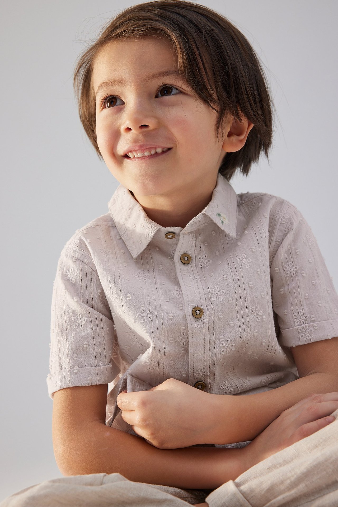 Framifrån: leende barn i ljuslila kortärmad bomullsskjorta med vit brodyr, knappar och ljusa byxor.