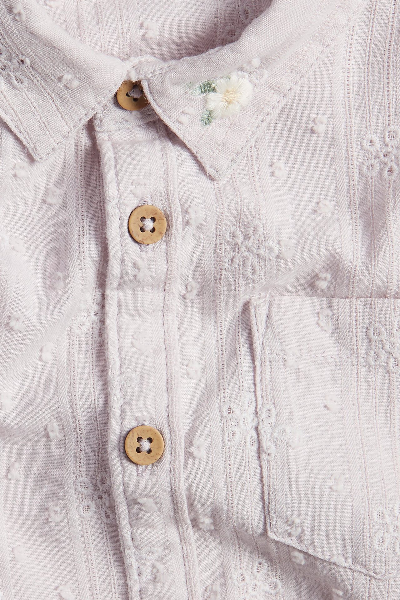 Zbliżenie na jasnofioletową bawełnianą koszulę dziecięcą z krótkim rękawem, kropkami, drewnianymi guzikami i haftem na kołnierzu.