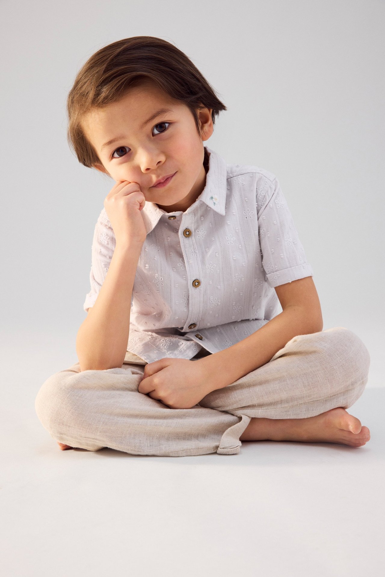 Widok z przodu: Dziecko w jasnoliliowej, haftowanej koszuli z krótkim rękawem, brązowymi guzikami i beżowych spodniach.