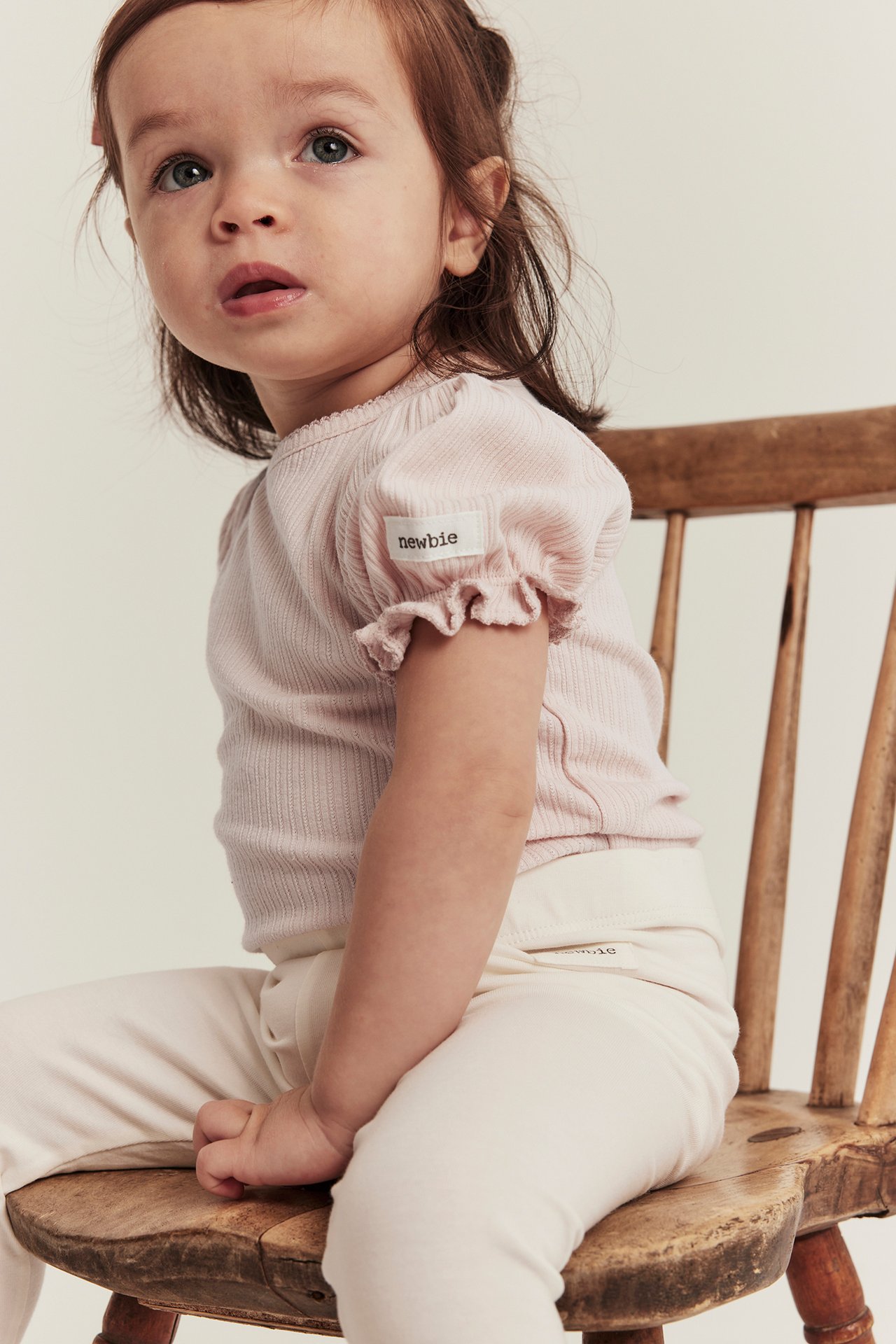 Sidovy av en babyflicka i ljusrosa ribbad body med puffärm och ljusbeige byxor.