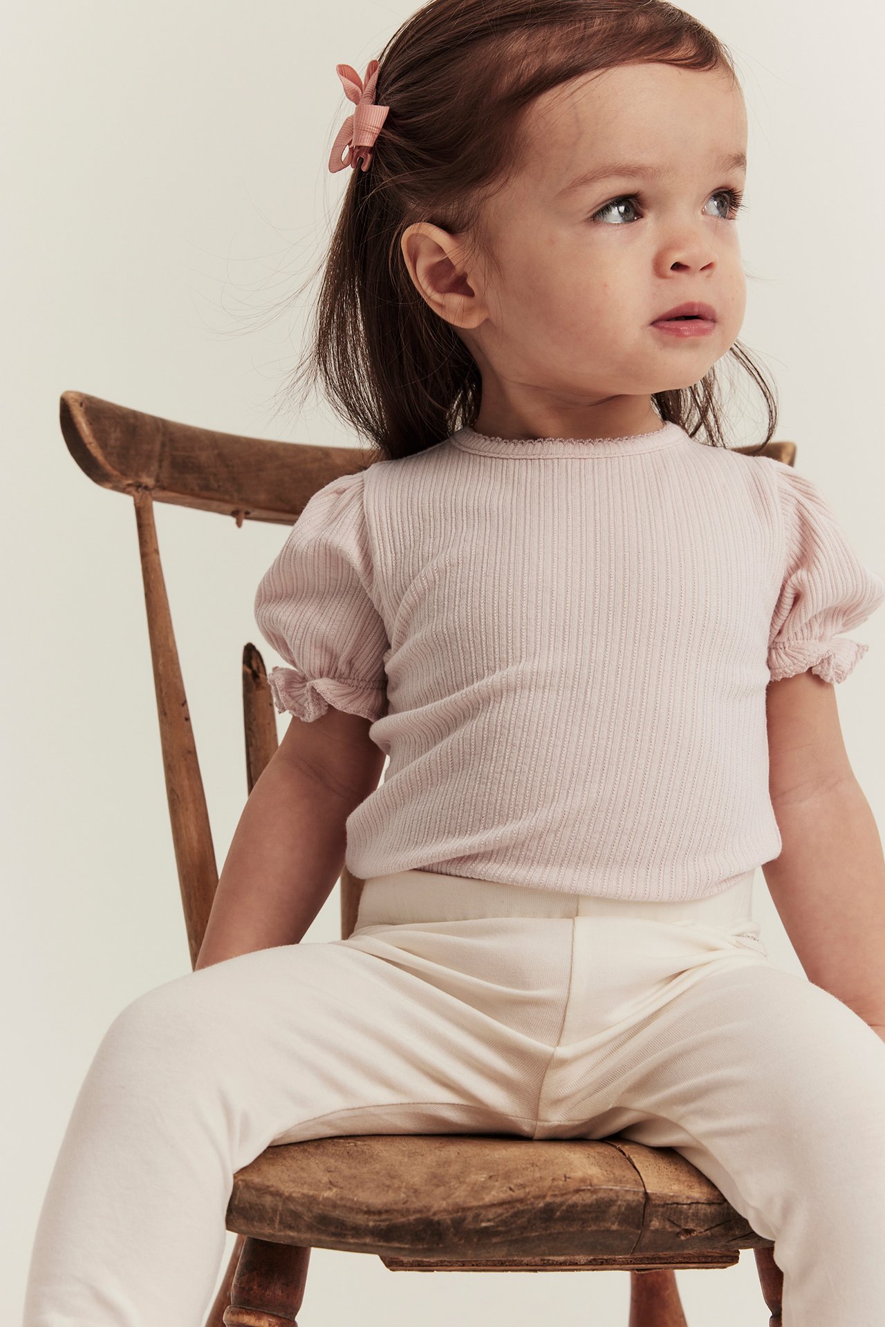 Frontvy av en baby i ljusrosa ribbad jerseybody med puffärm, gräddvita leggings och rosa hårklämma.