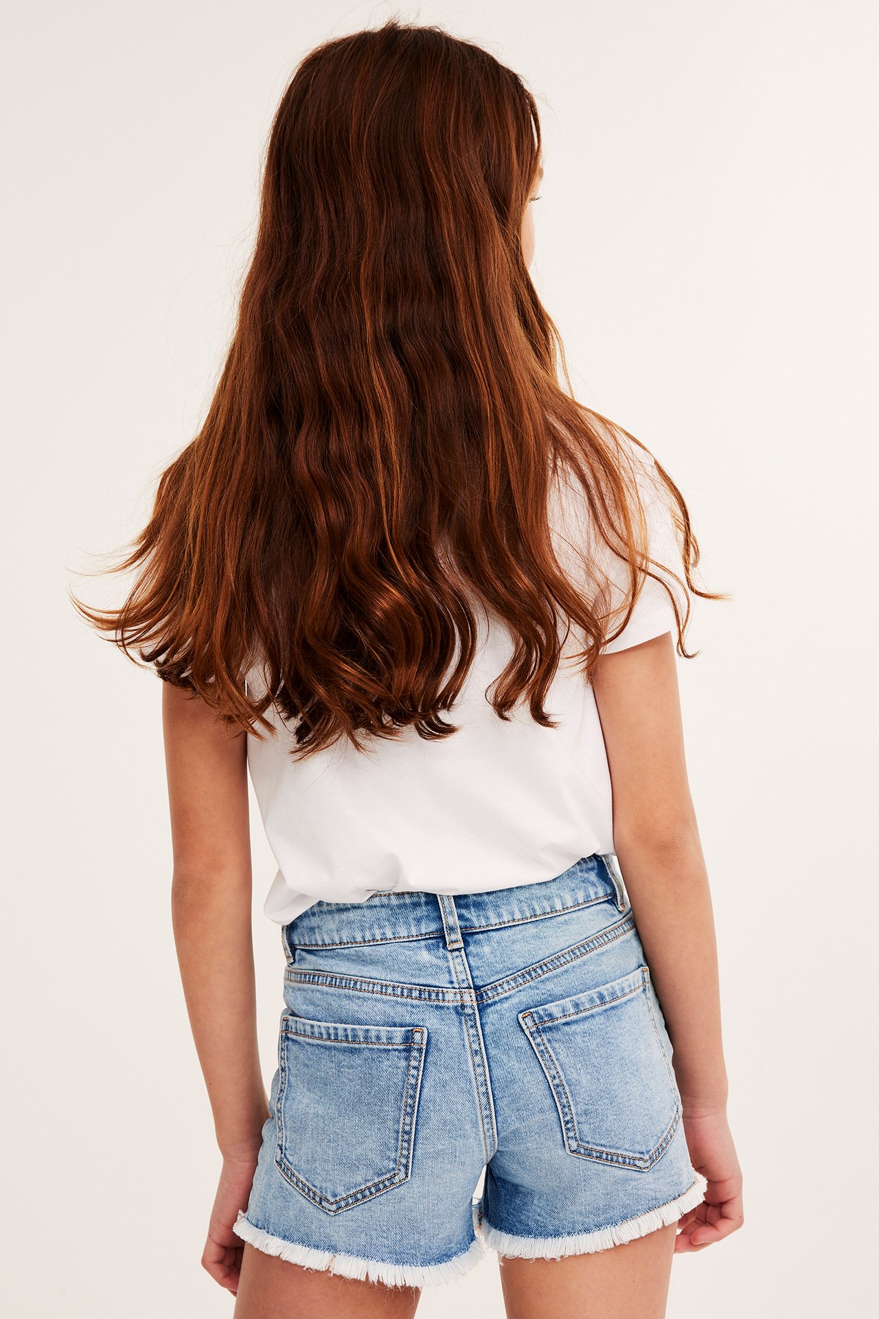 Bakifrån: ett barn i ljusa jeansshorts med rå kant och en vit t-shirt.
