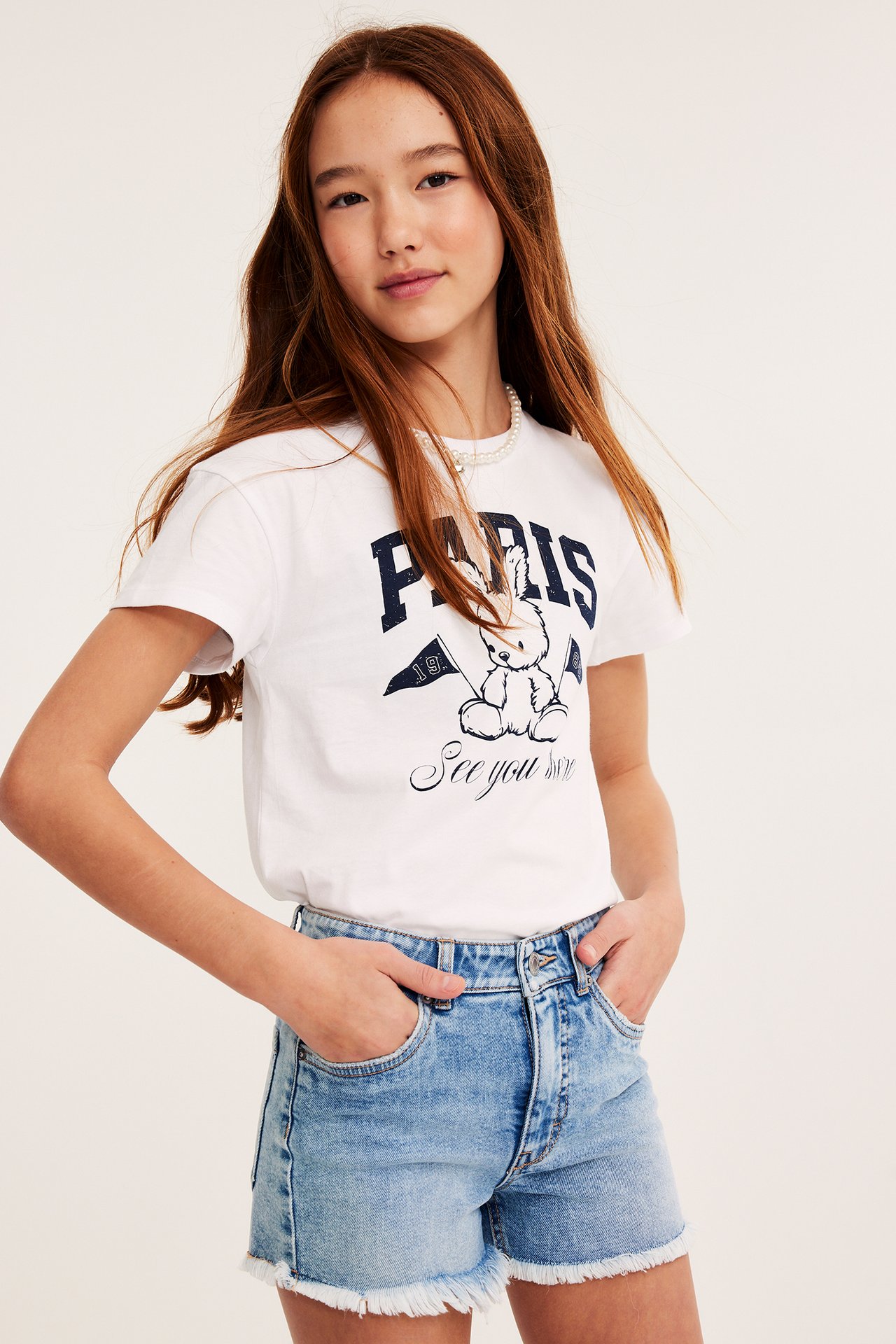 Barn i ljusblå jeansshorts med rå kant, vit t-shirt med Paris-tryck och pärlhalsband.