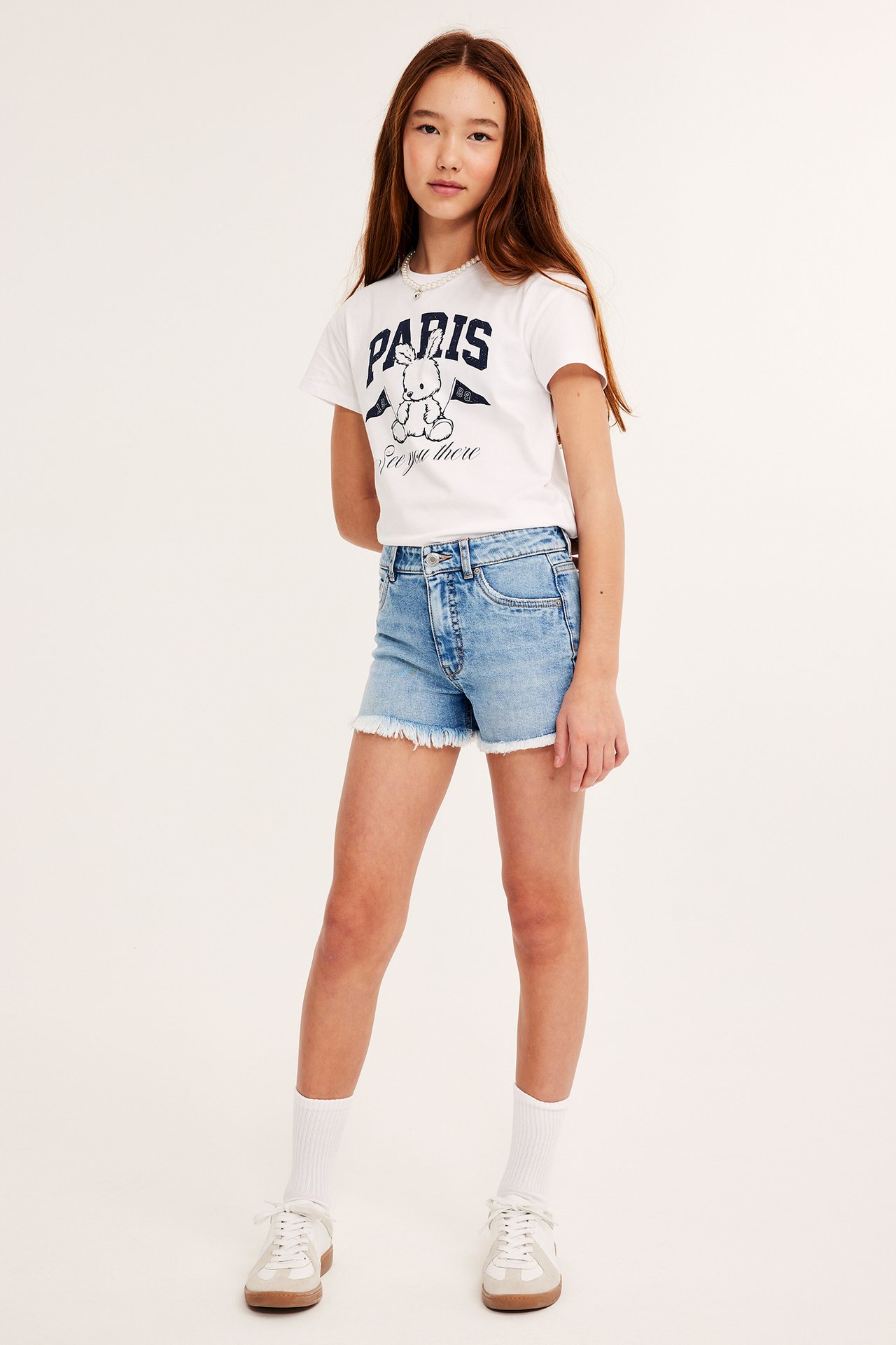 Frontvy: Barn i ljusblå denimshorts med råa kanter, vit grafisk t-shirt och vita sneakers.