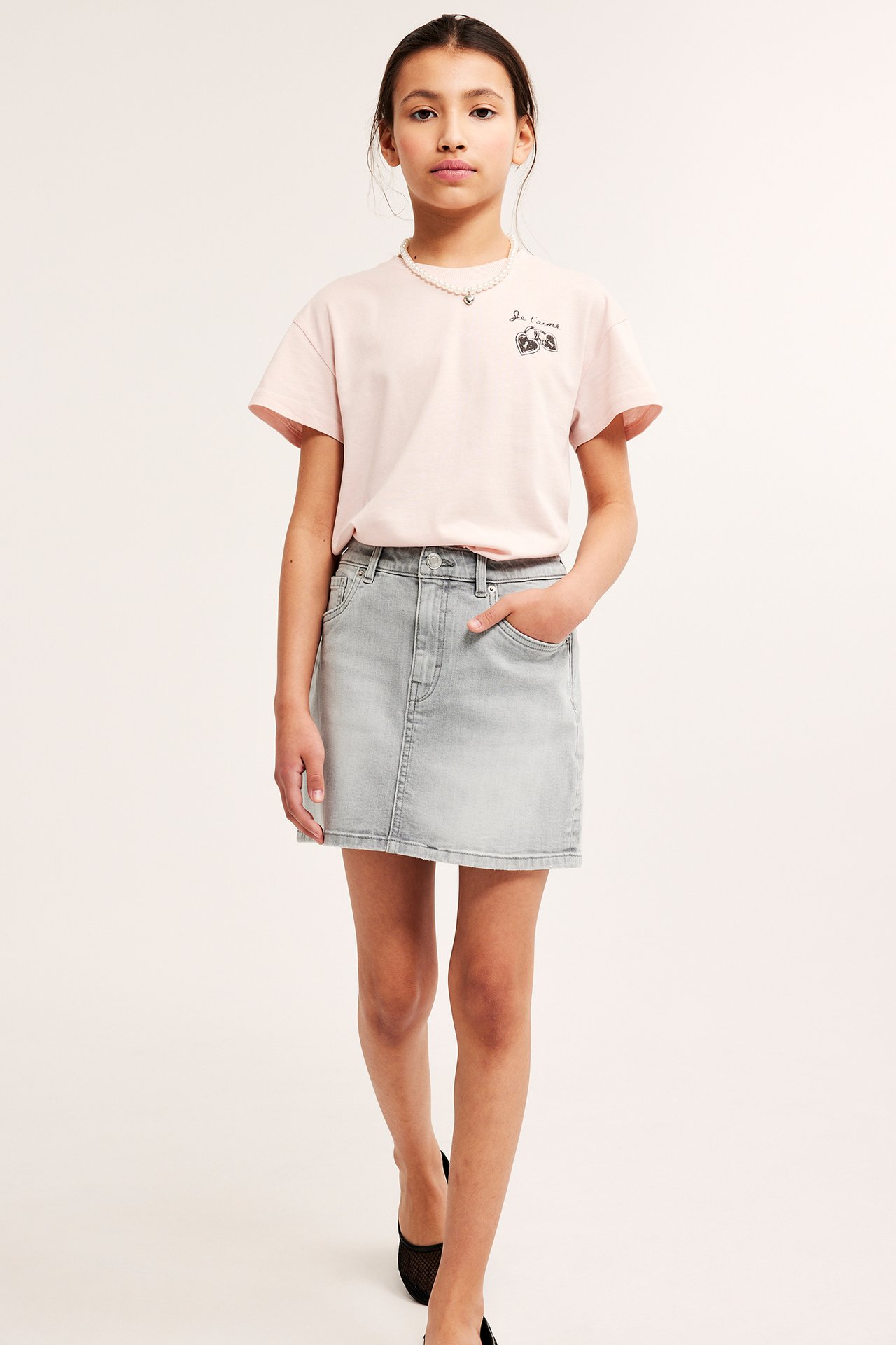 Frontvy av ett barn i en ljusgrå jeanskjol, rosa t-shirt med tryck, pärlhalsband och svarta skor.