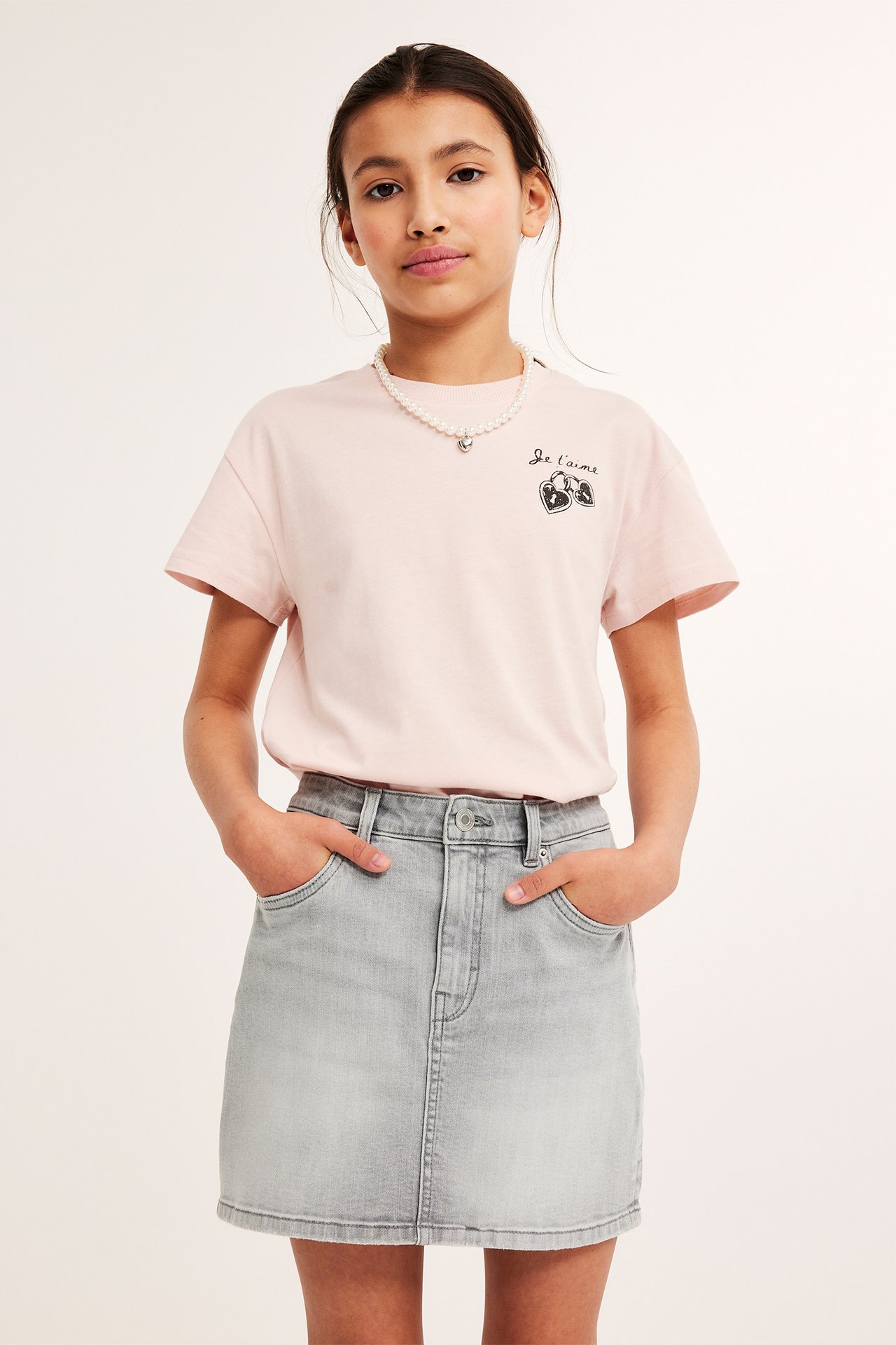 Framsida av ett barn i kort grå jeanskjol, ljusrosa tryckt t-shirt och pärlhalsband.