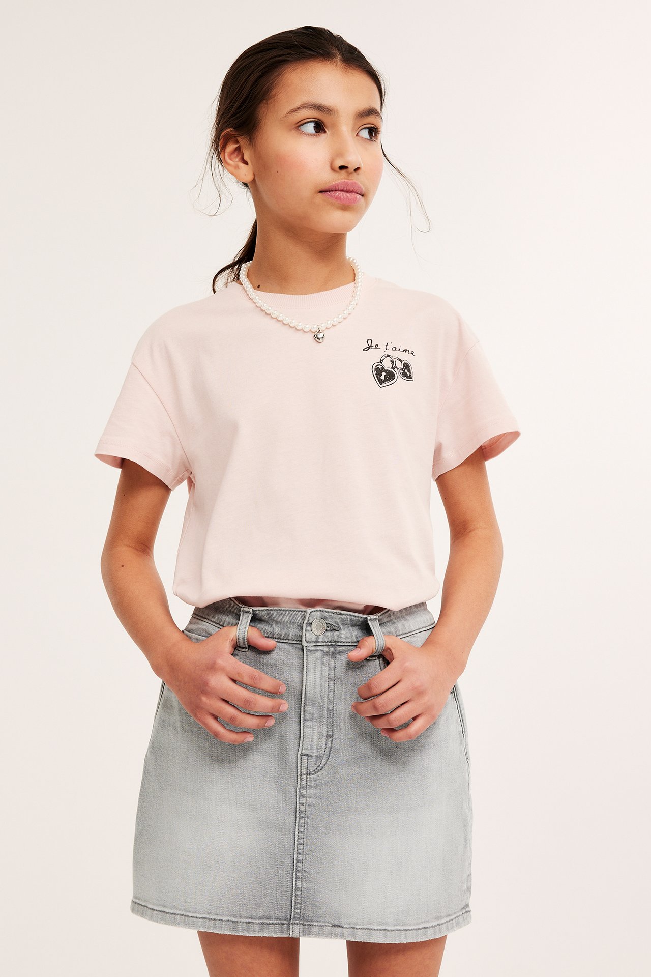 Framifrån. Ett barn klädd i rosa t-shirt med tryck vid bröstet. Barnet har händerna hängandes framtill i hällorna på den gråa kjolen i denim. Barnet bär ett halsband och har mörkt hår uppsatt i en toffs.