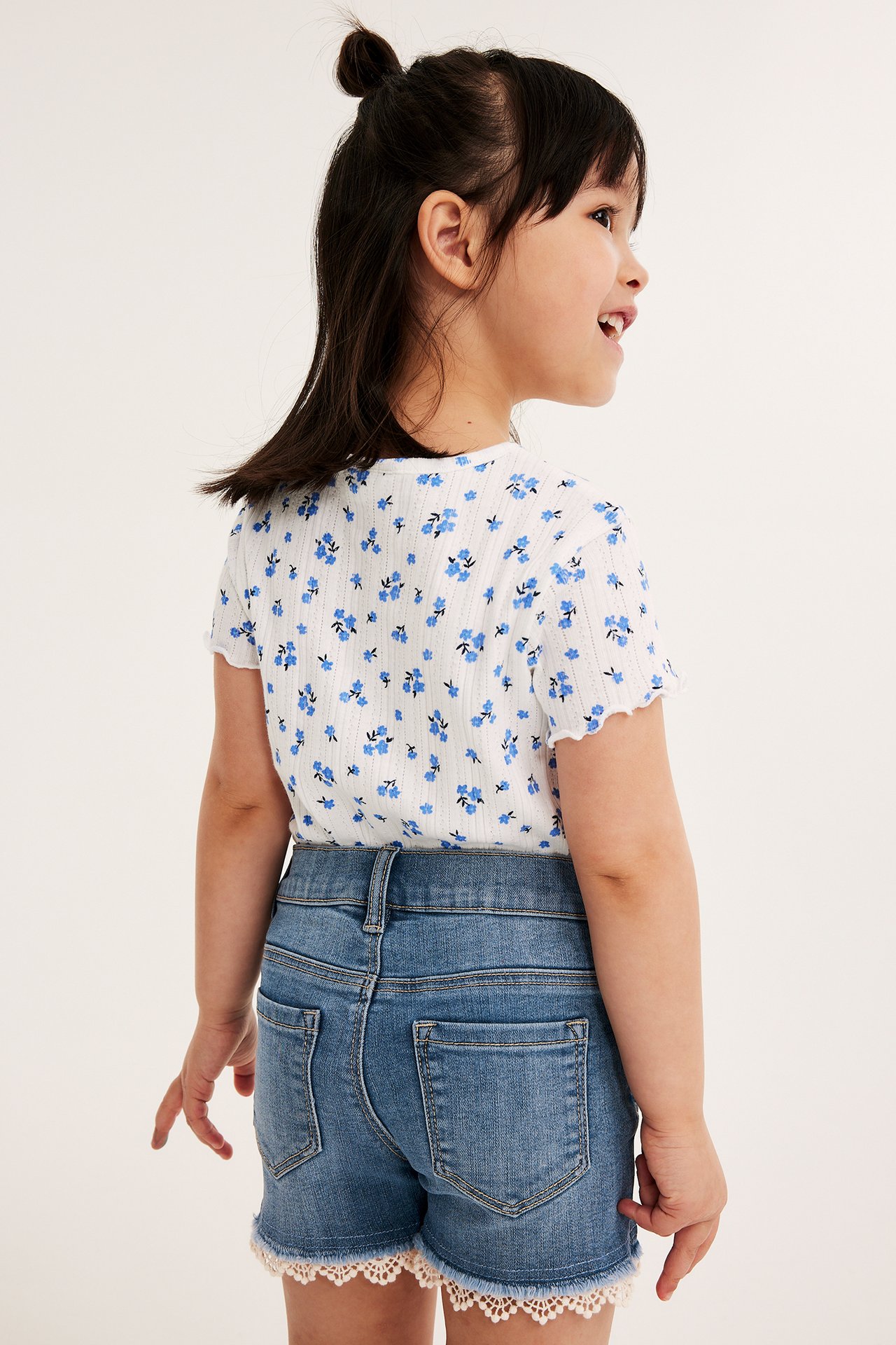 Rygg-/sidovy av ett barn i ljusa jeansshorts med vit spetskant och en vit blommig t-shirt.