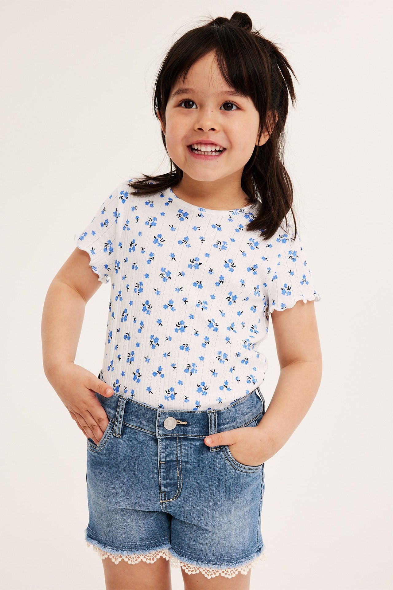 Widok z przodu: Dziecko w jasnych szortach jeansowych z białą koronką i białym, kwiecistym T-shirtem w prążki.