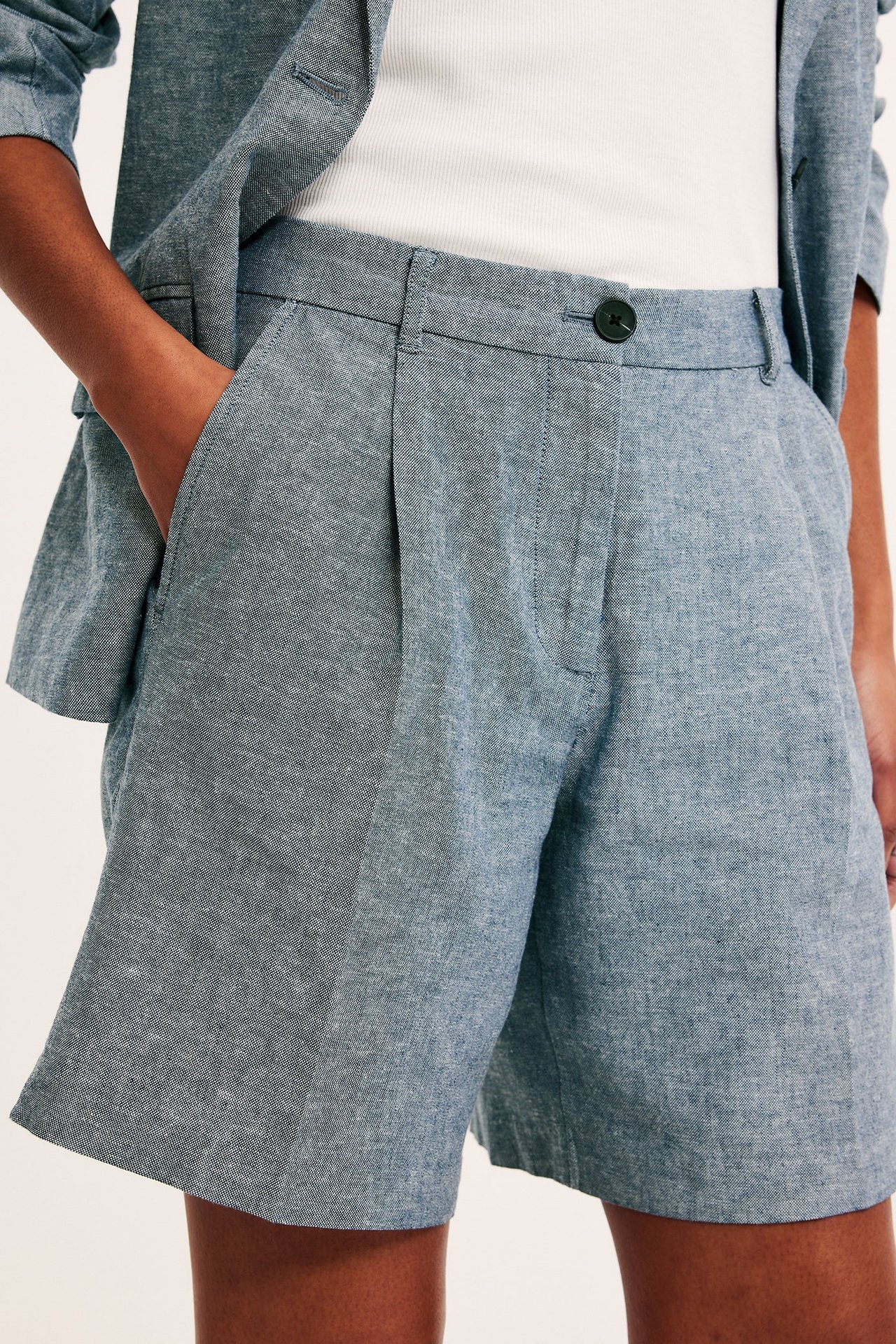 Lähikuva edestä: nainen sinisissä meleeratuissa pellavashortseissa, taskut, valkoinen ribbitoppi ja bleiseri.