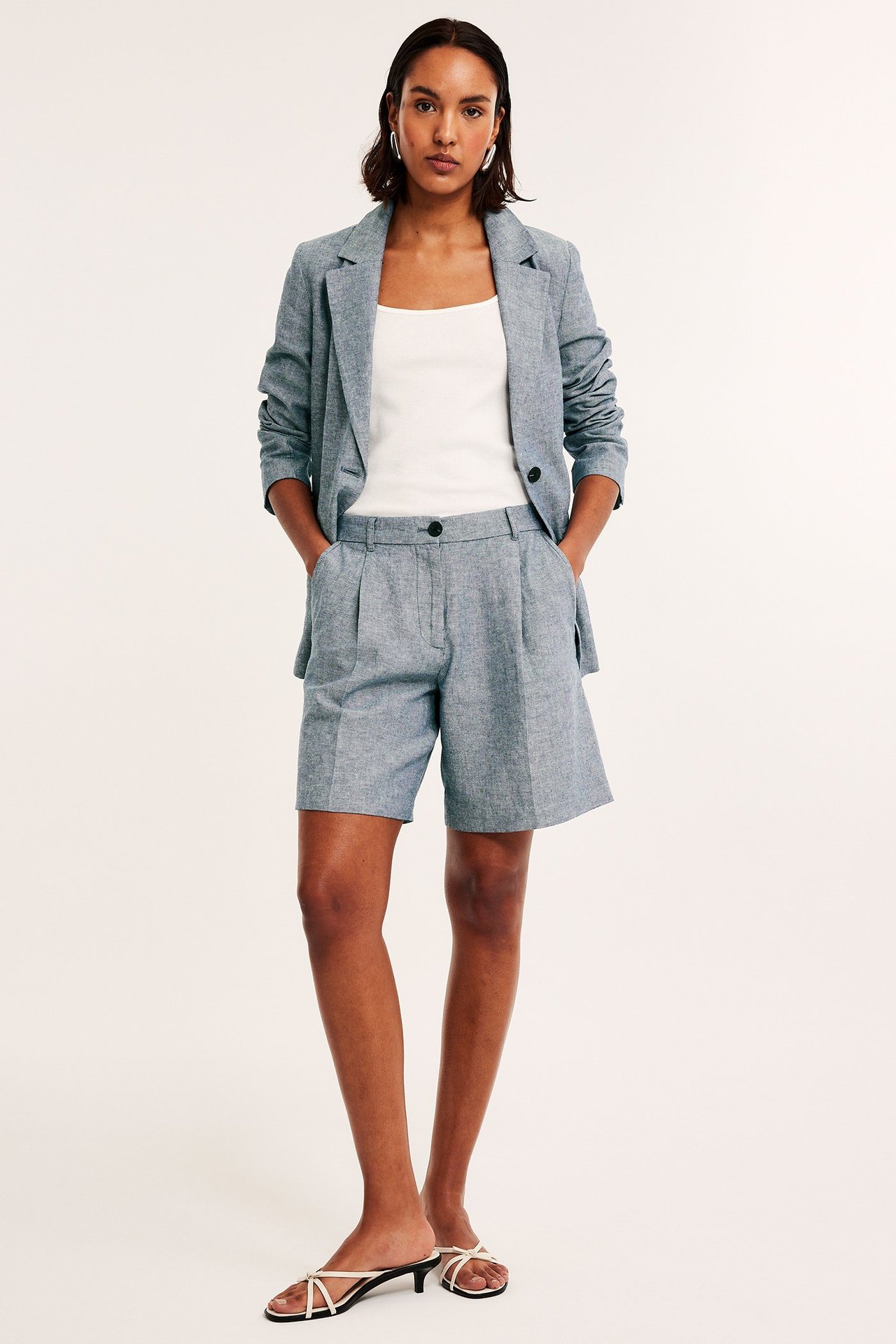 Edestä: Nainen sinimelange pellavashortseissa, bleiserissä, valkoisessa topissa ja valkoisissa sandaaleissa.