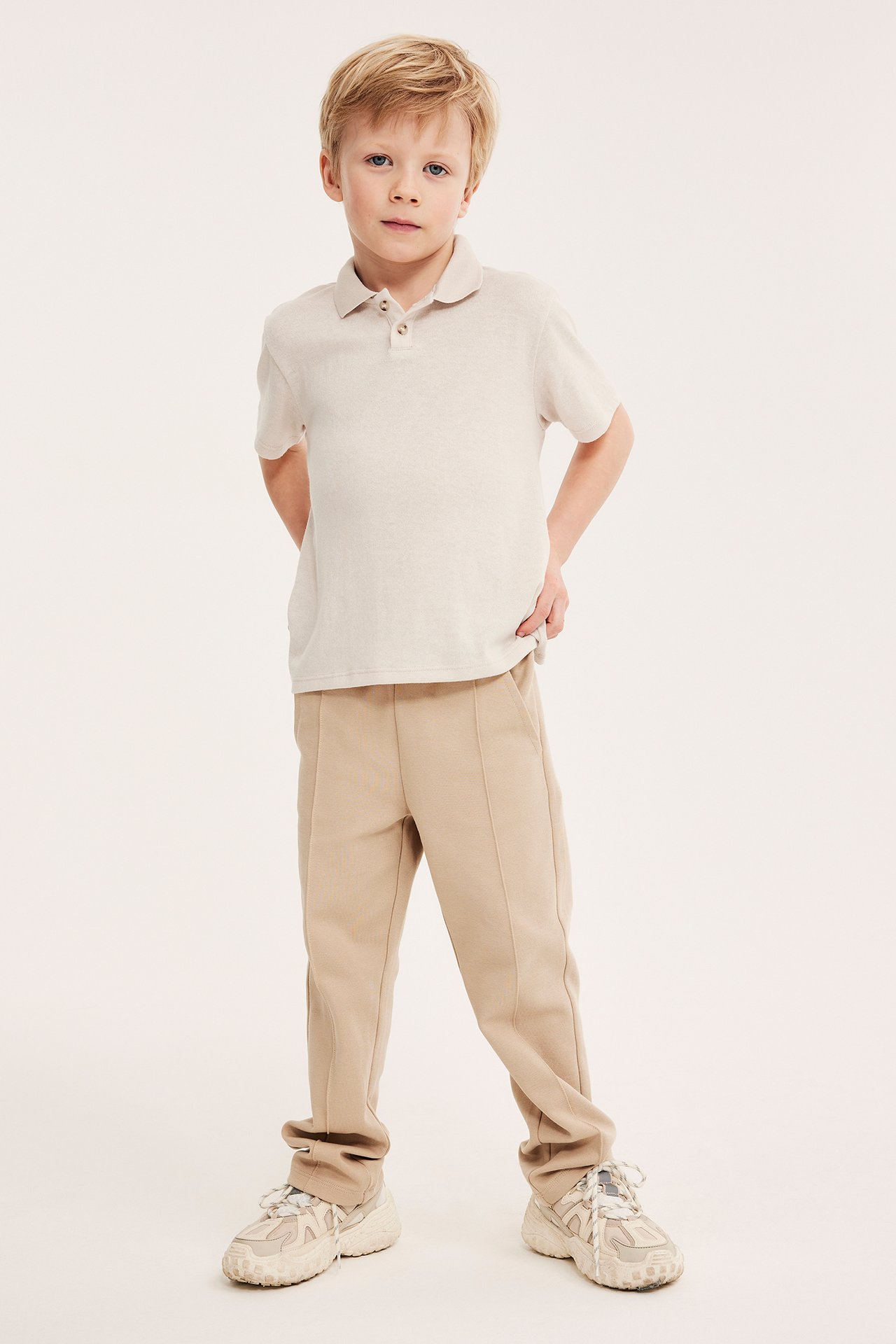 Ett barn bär en ljusbeige finstickad kortärmad pikétröja, beige byxor och ljusa sneakers.