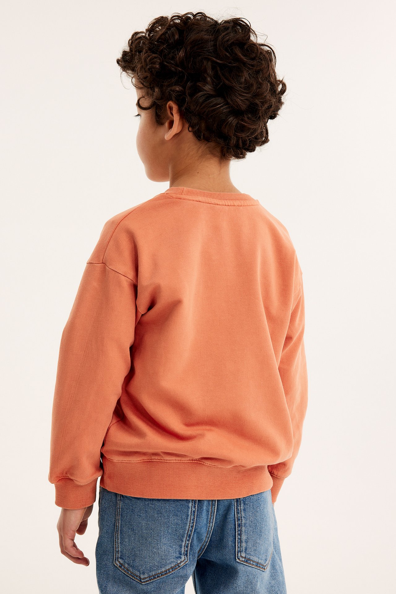 Bakifrån, ett barn i en orange tvättad bomullssweatshirt med ribbade muddar och blå jeans.