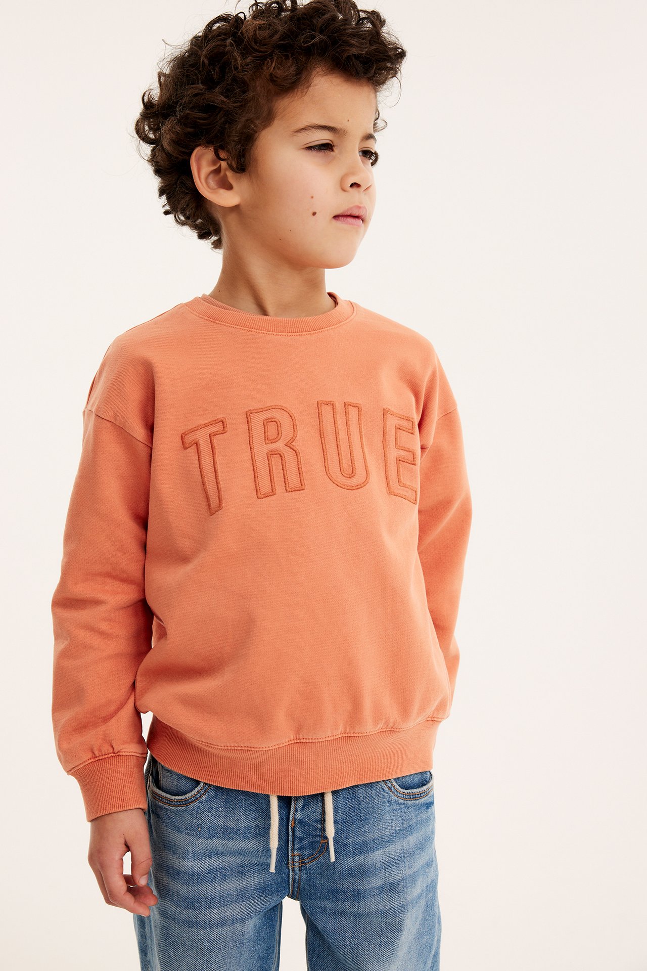 Framifrån, ett barn i en orange bomullssweatshirt med 'TRUE'-broderi, stylad med blå jeans.