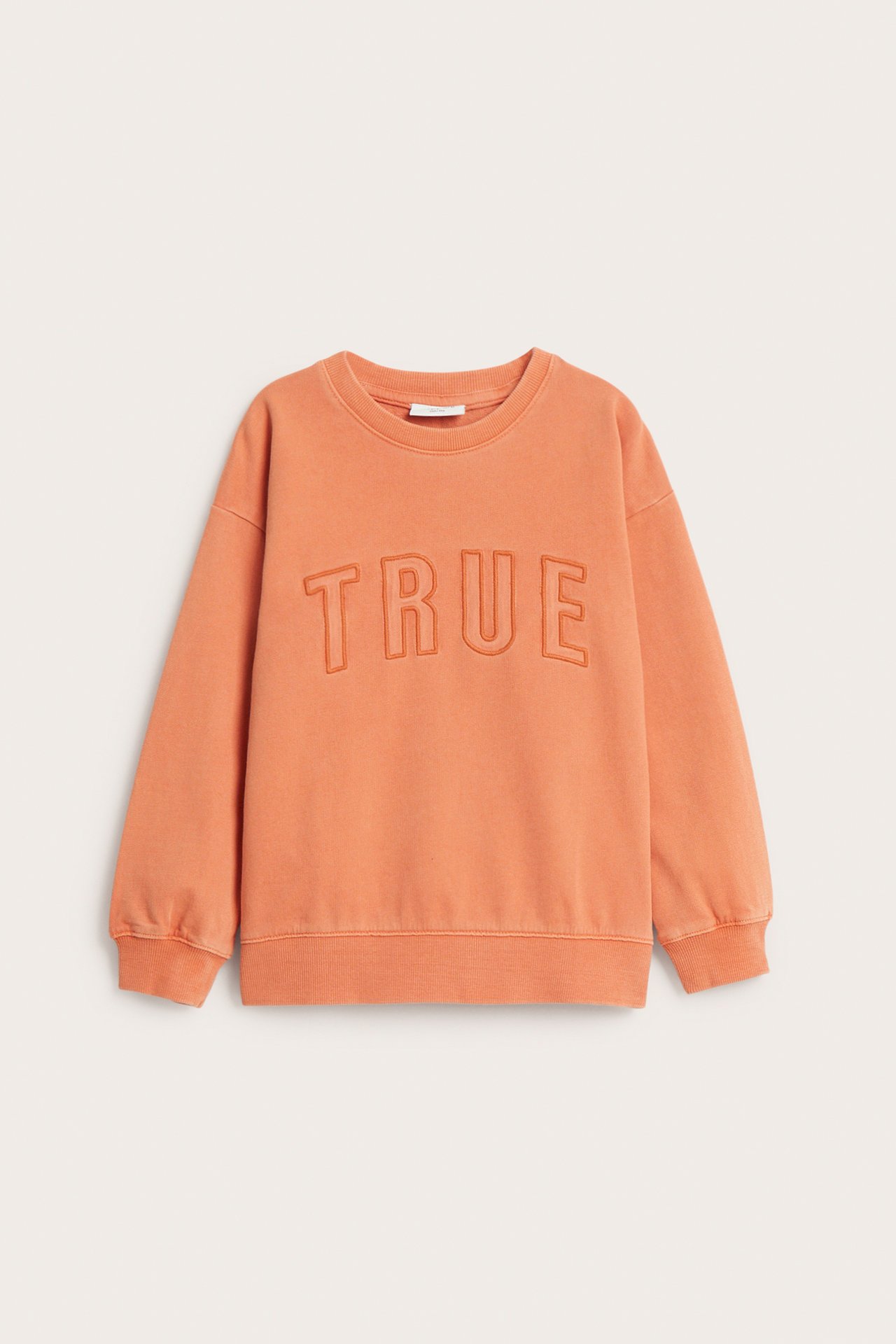 Frontvy av en orange bomullssweatshirt med tvättad look och "TRUE" broderat framtill.