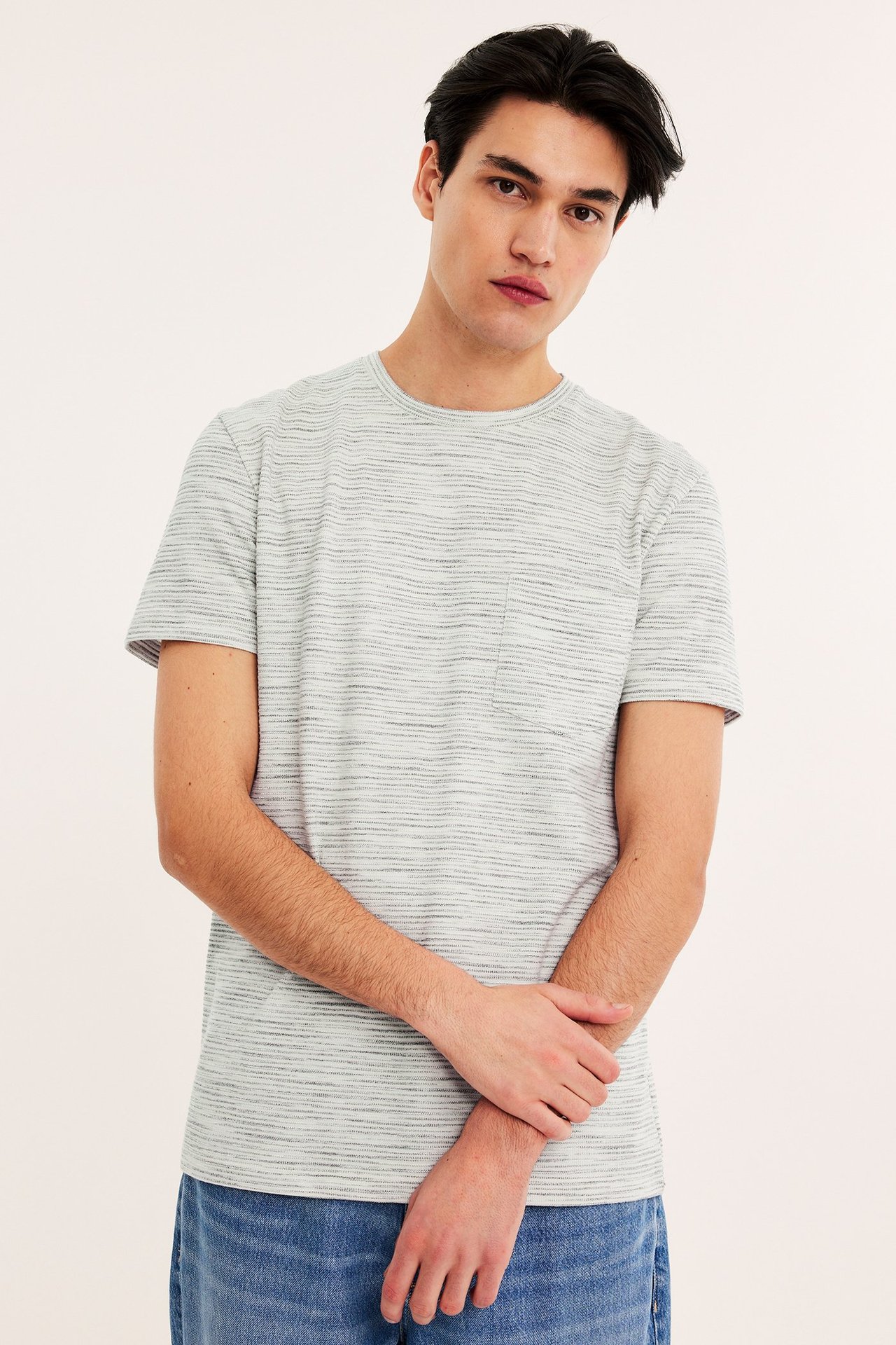 Edestä: Mies yllään luonnonvalkoinen strukturoitu meleerattu t-paita rintataskulla ja siniset farkut.