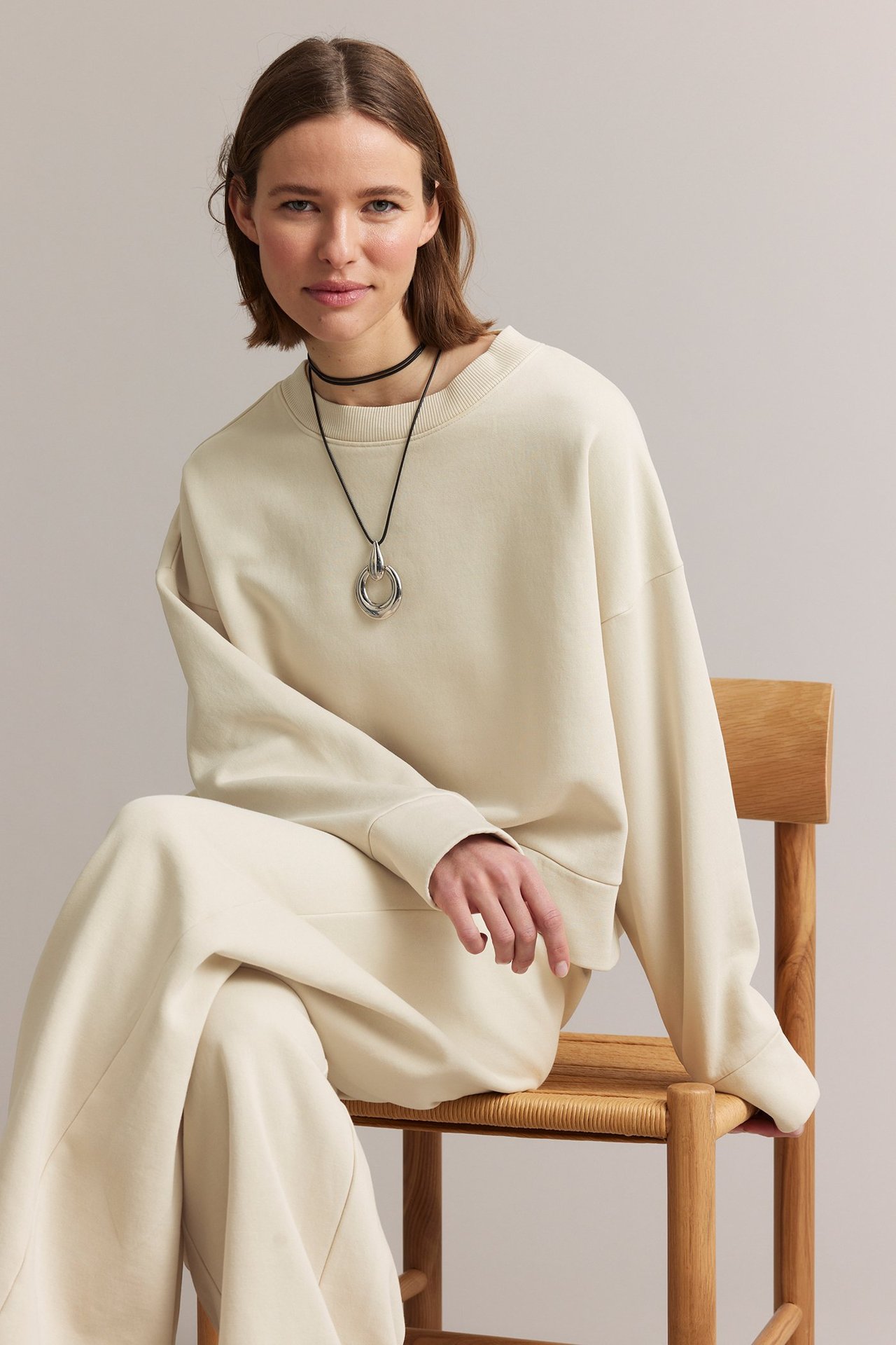 Frontvy av en kvinna i en ljusbeige jerseyströja, matchande vida byxor och ett svart-silverfärgat halsband.