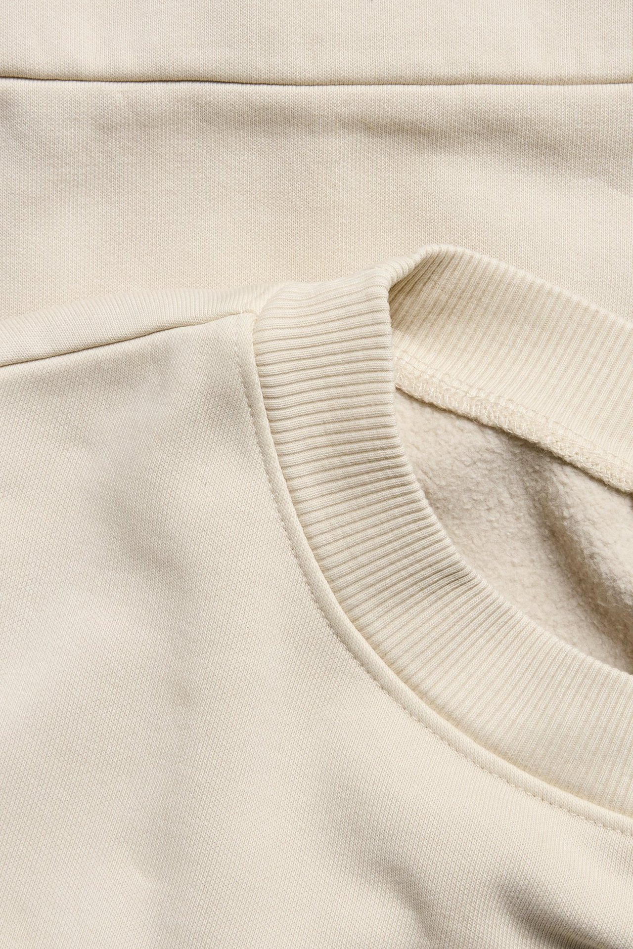 Zbliżenie na jasnobeżową bluzę dresową z dzianiny z prążkowanym okrągłym dekoltem i efektem sprania.