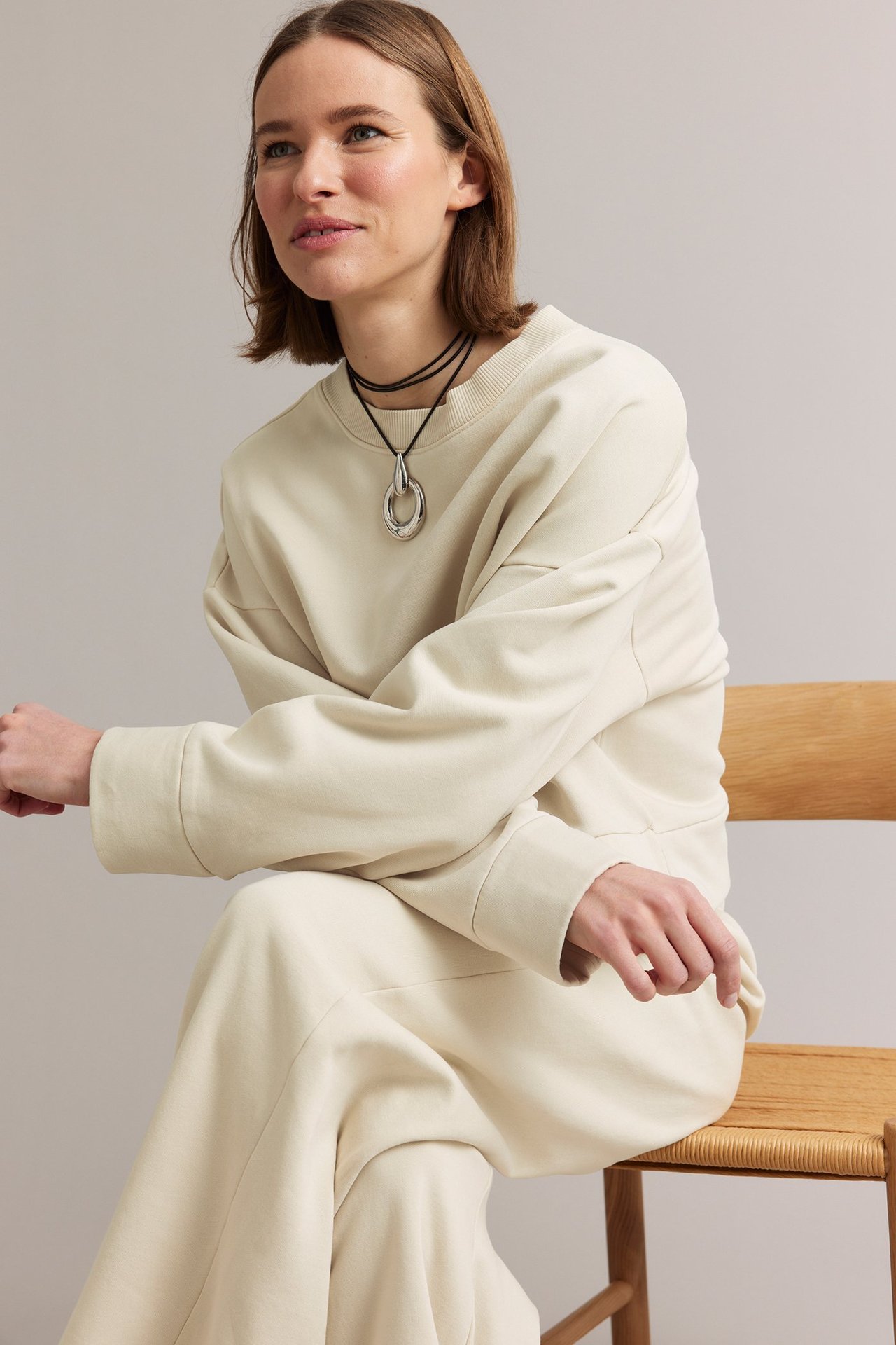Widok z boku na kobietę w jasnobeżowej bluzie dresowej z dżerseju, pasujących spodniach i srebrnym naszyjniku.