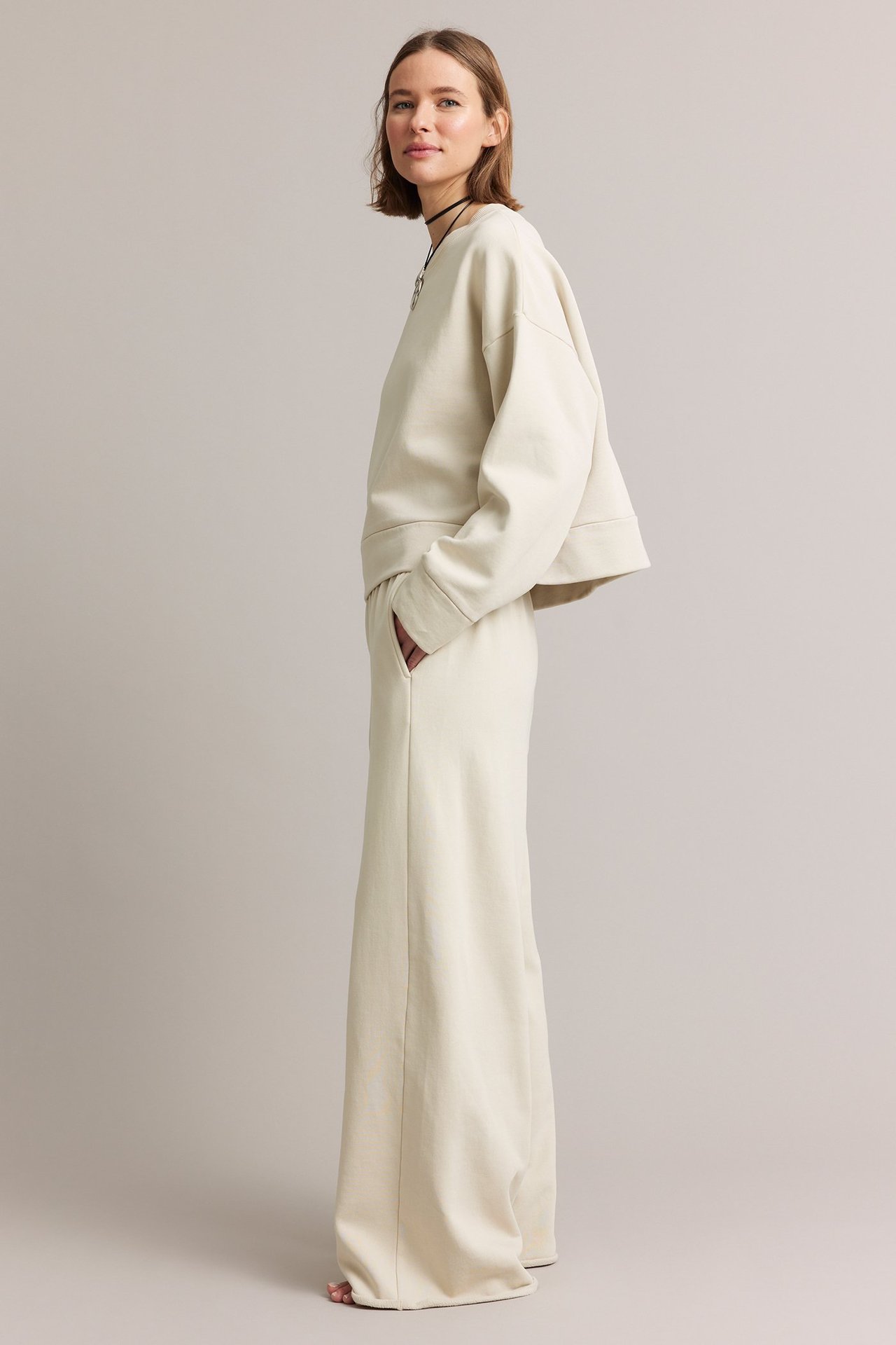 Sidebilde av en kvinne i lys beige rette jersey-joggebukser med lommer og en matchende kort genser.