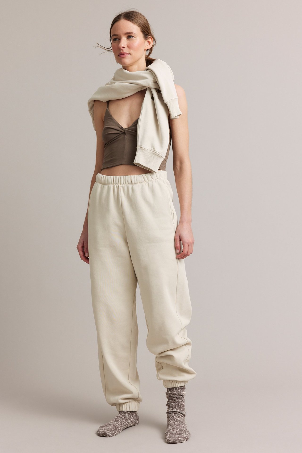 Frontutsikt: Kvinne i lysebeige joggebukse, en brun crop top og en lysebeige genser drapert.