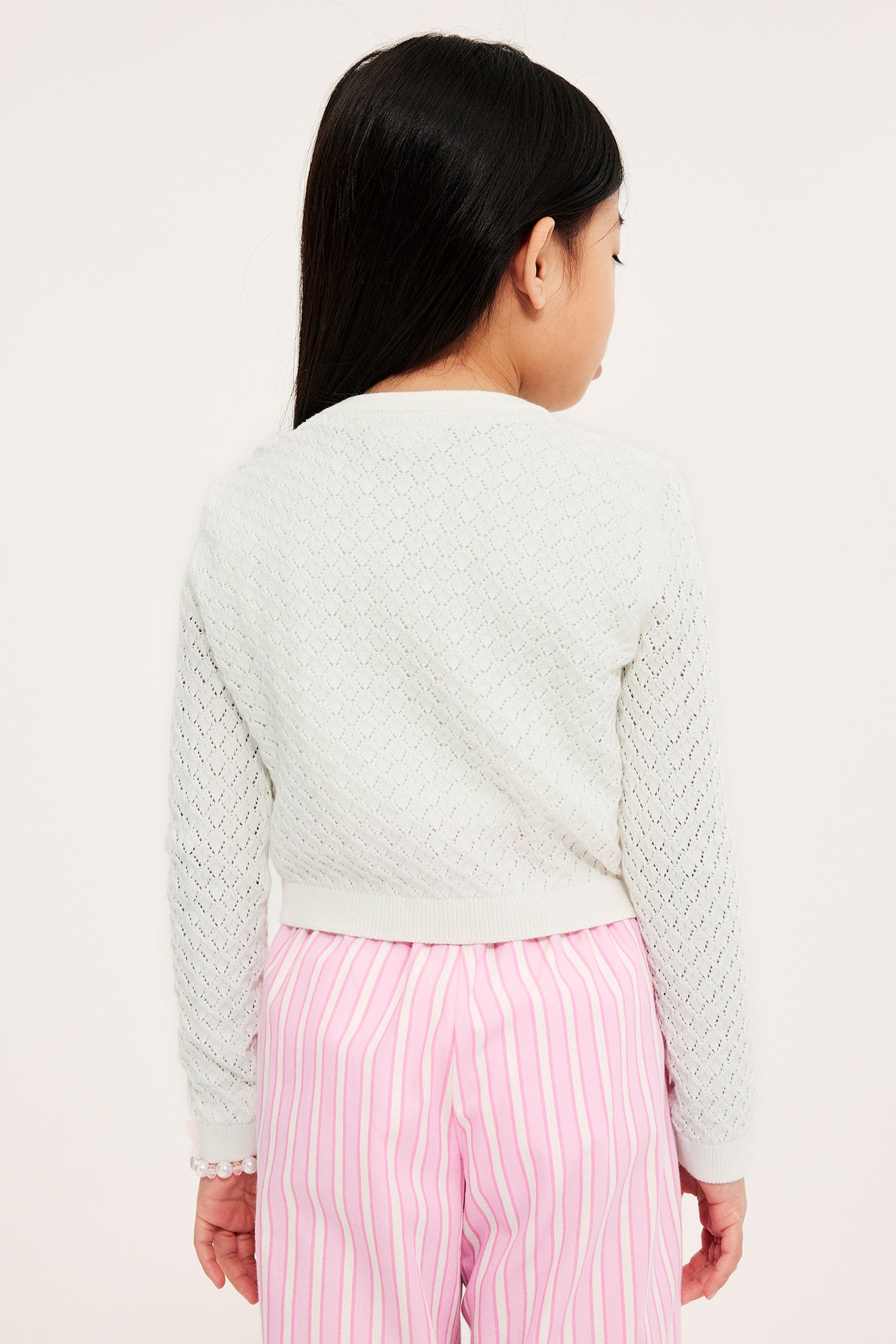 Ryggvisning av et barn i en offwhite rundhalset hullmønstret cardigan over rosa- og hvitstripete bukser.