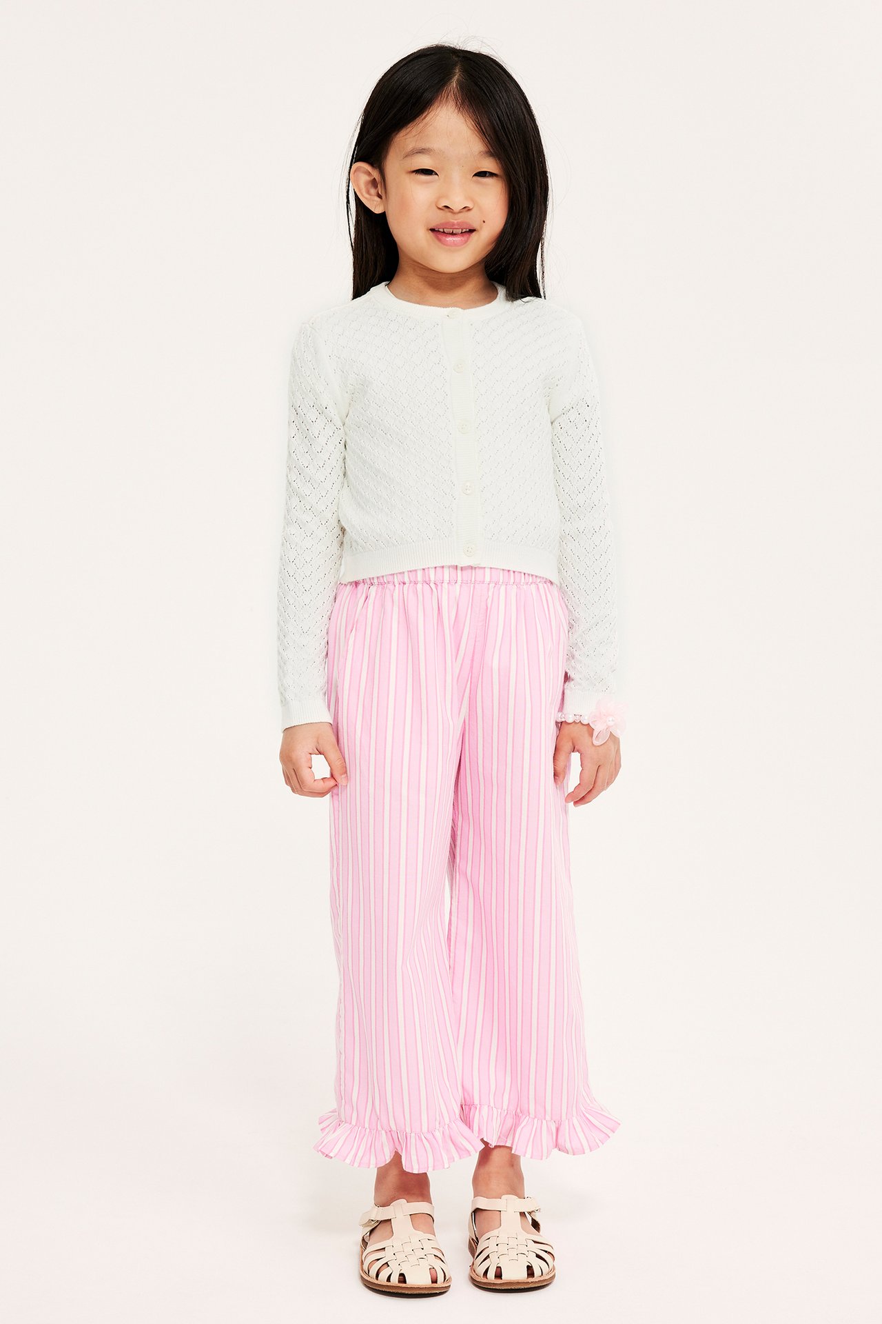 Forfra: et barn i offwhite hullmønstret cardigan, rosa stripete bukser og beige sandaler.