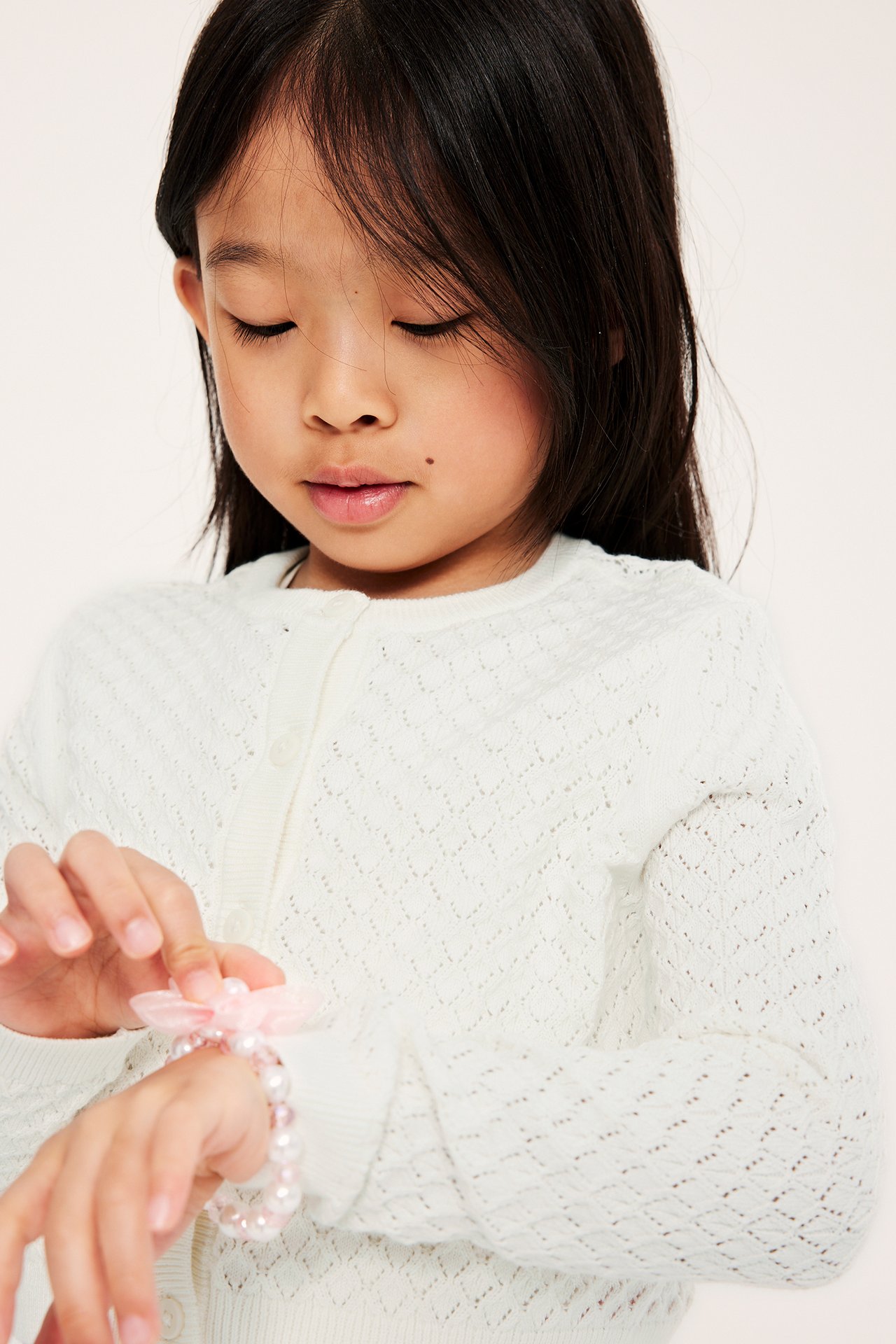 Nærbilde forfra av et barn i offwhite hullmønstret cardigan med knapper, justerer et rosa perlearmbånd.