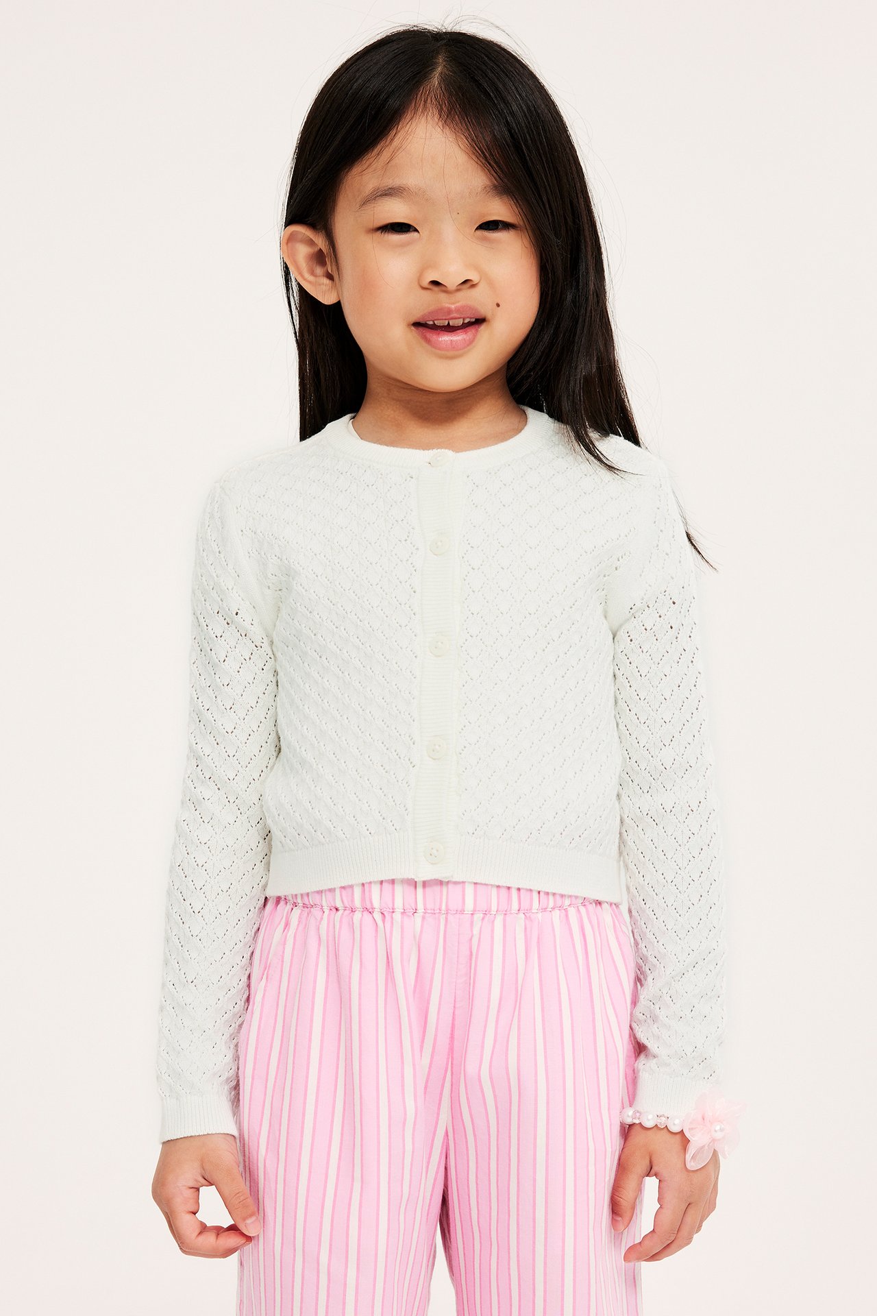 Frontbilde av et barn i offwhite mønsterstrikket cardigan med knapper og rosa stripete bukser.