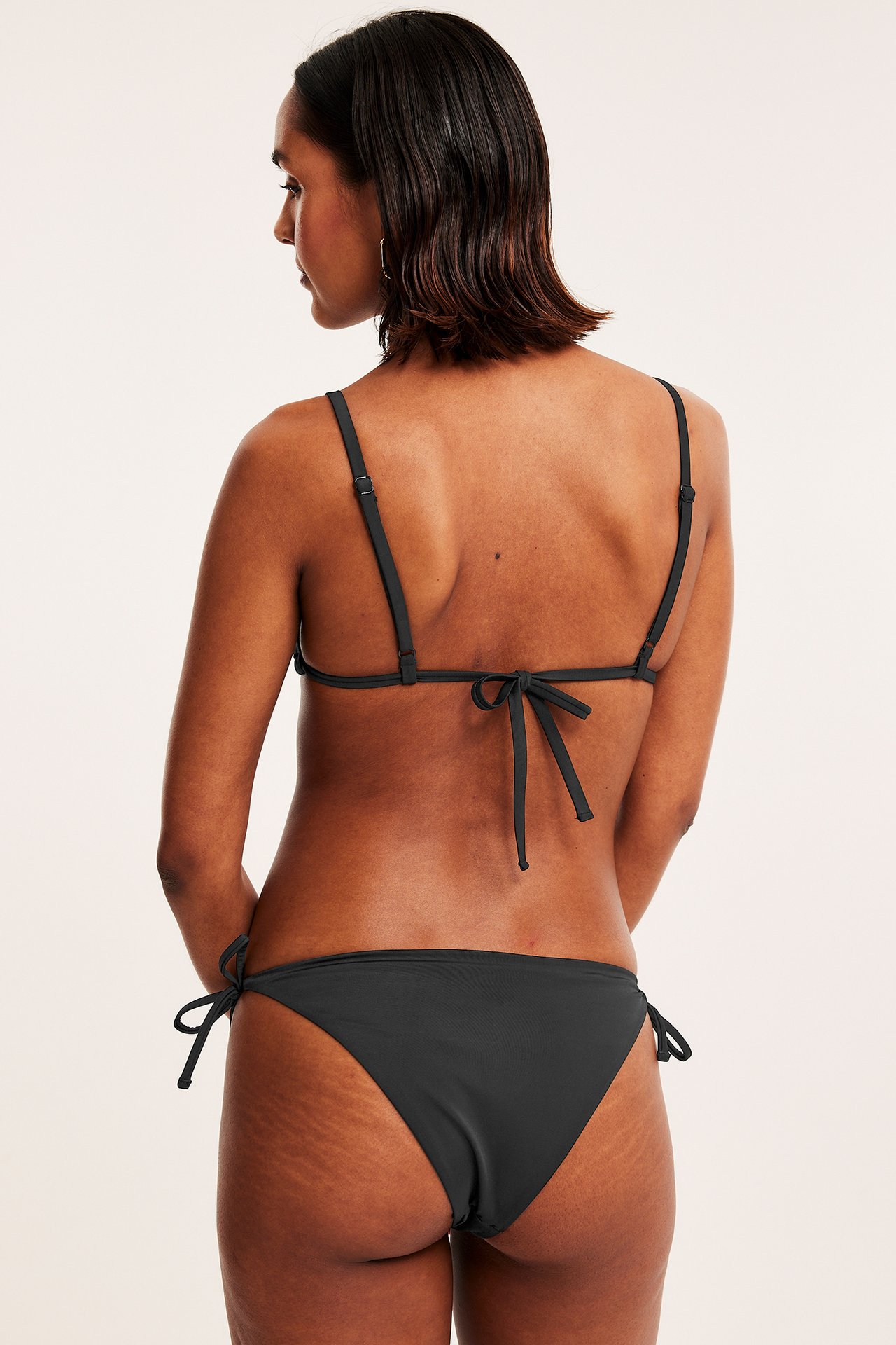 Bakifrån: kvinna i svart brazilian bikinitrosa med knytband och matchande svart bikinitopp.