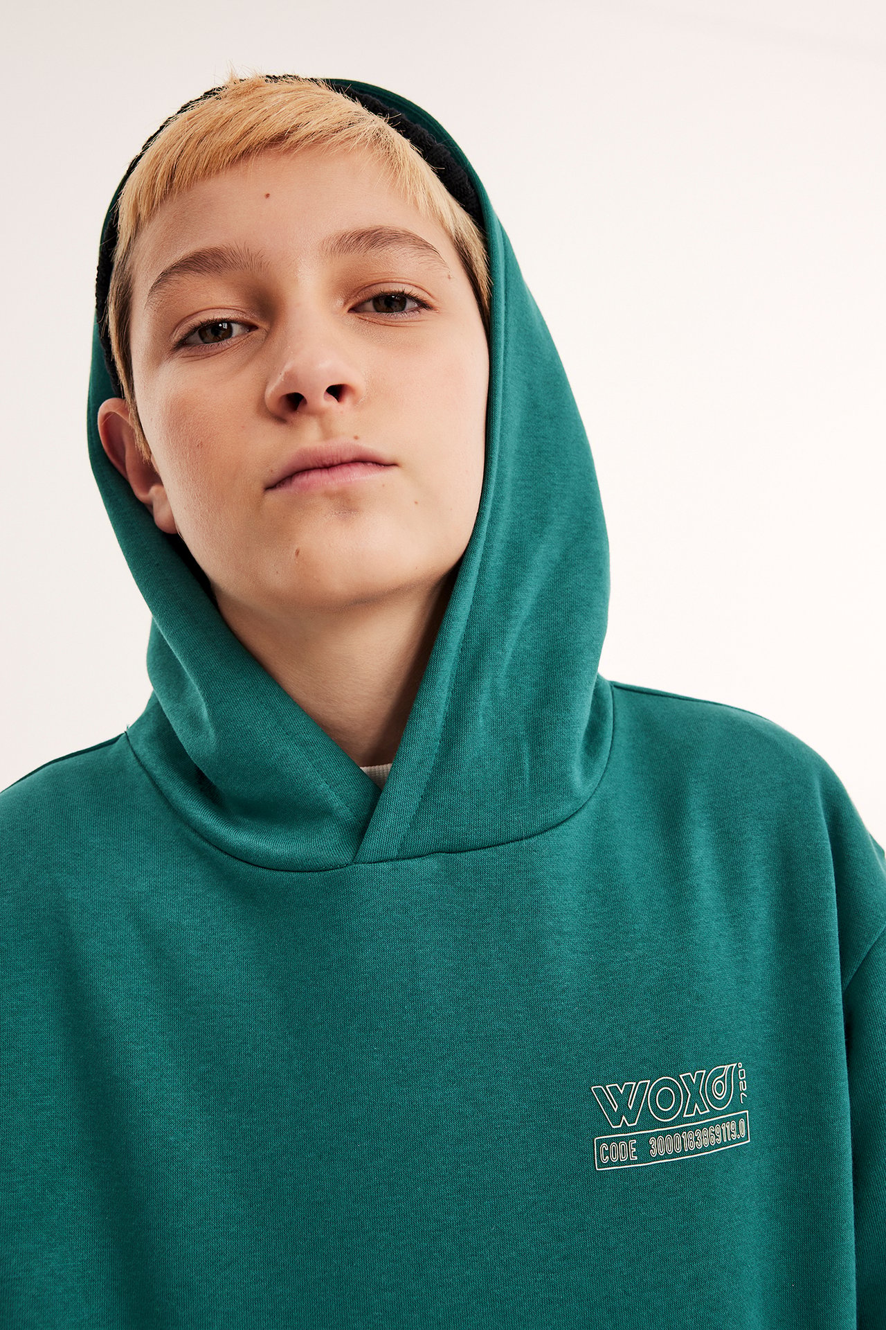 Nærbilde forfra av et barn i en mørkegrønn hoodie med hvitt brysttrykk og en svart lue.