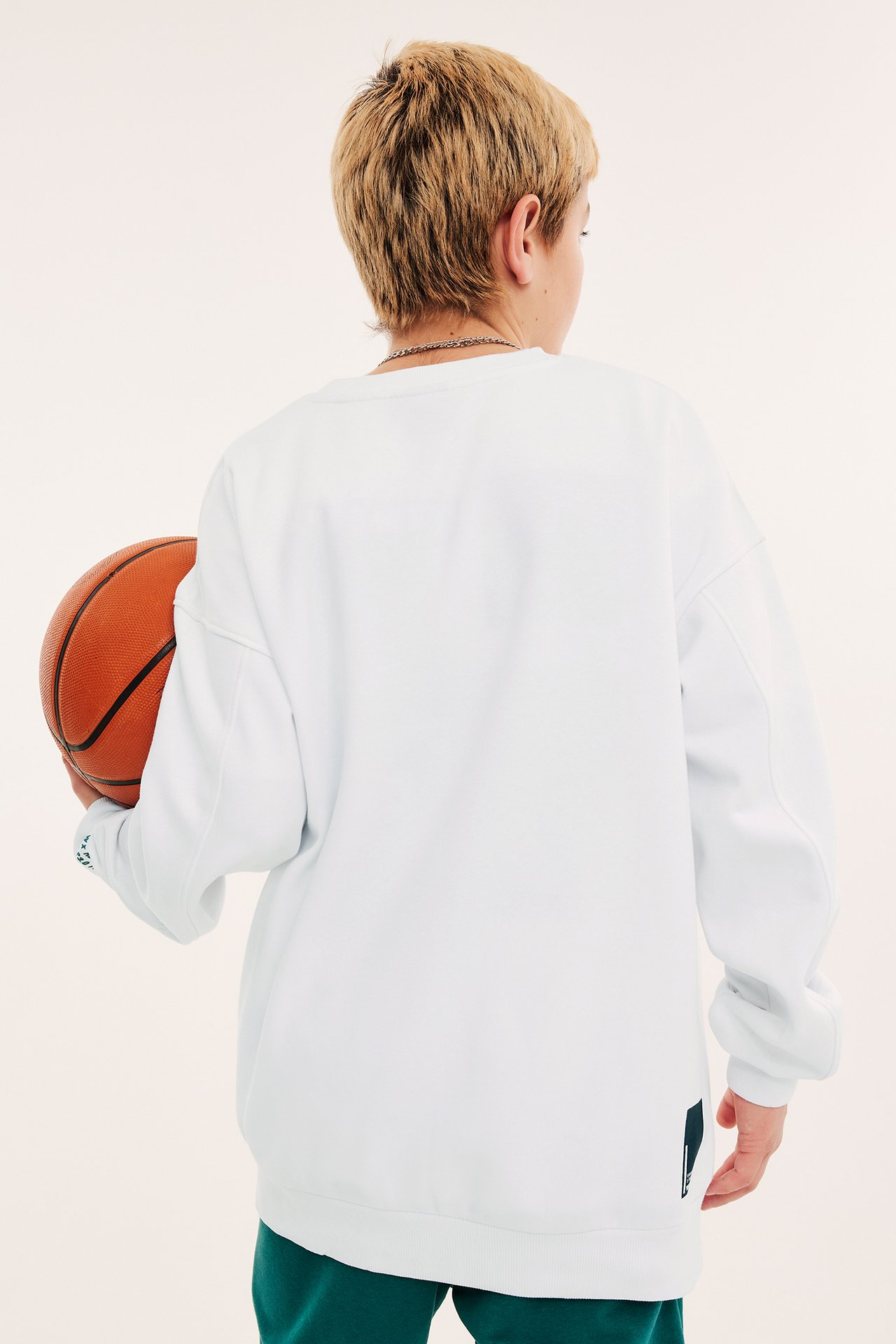 Bakifrån, ett barn i en vit lös sweatshirt med ribbade muddar, gröna byxor, håller en basketboll.