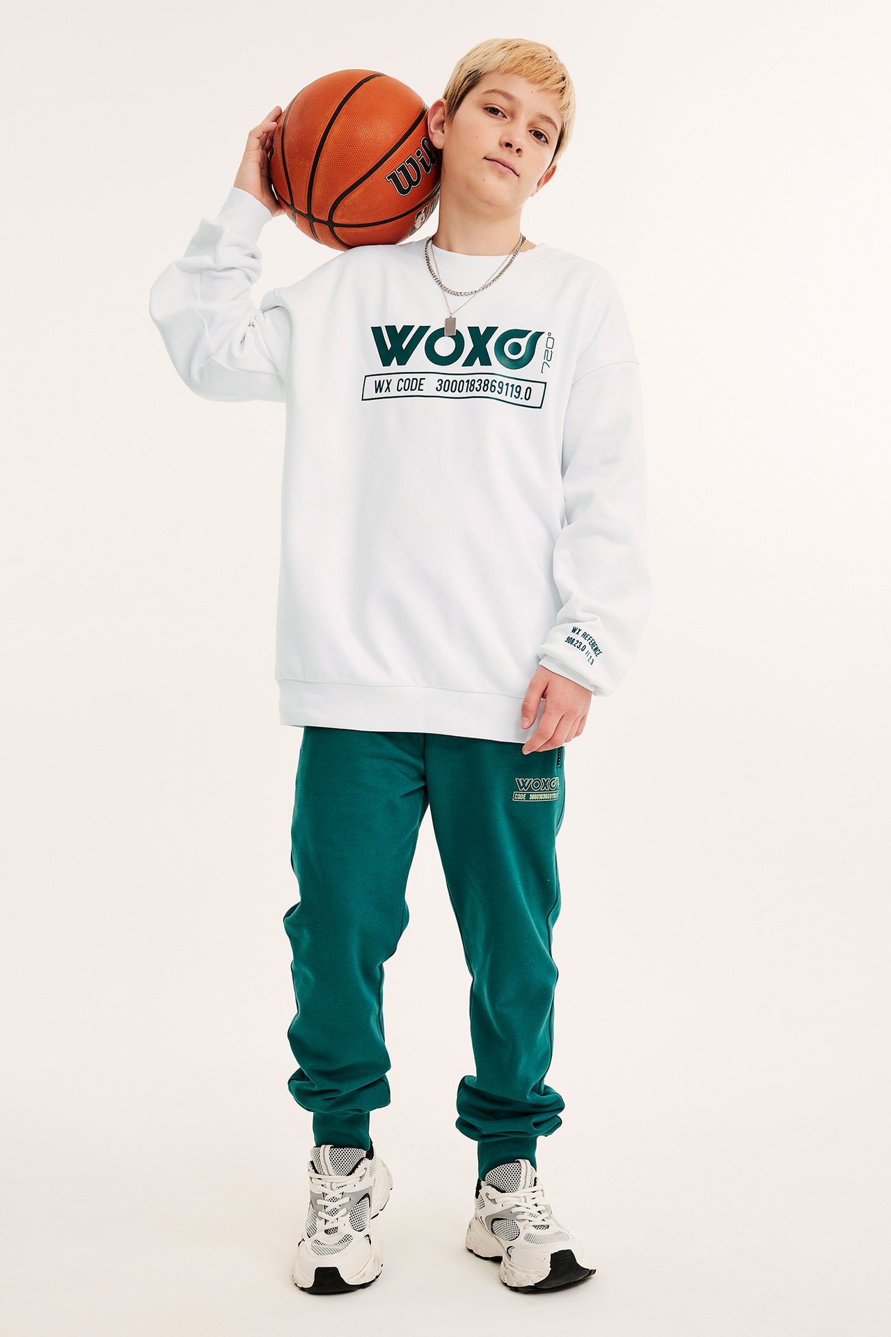 Frontvy av ett barn i vit mönstrad sweatshirt, gröna mjukisbyxor och sneakers, håller en basketboll.