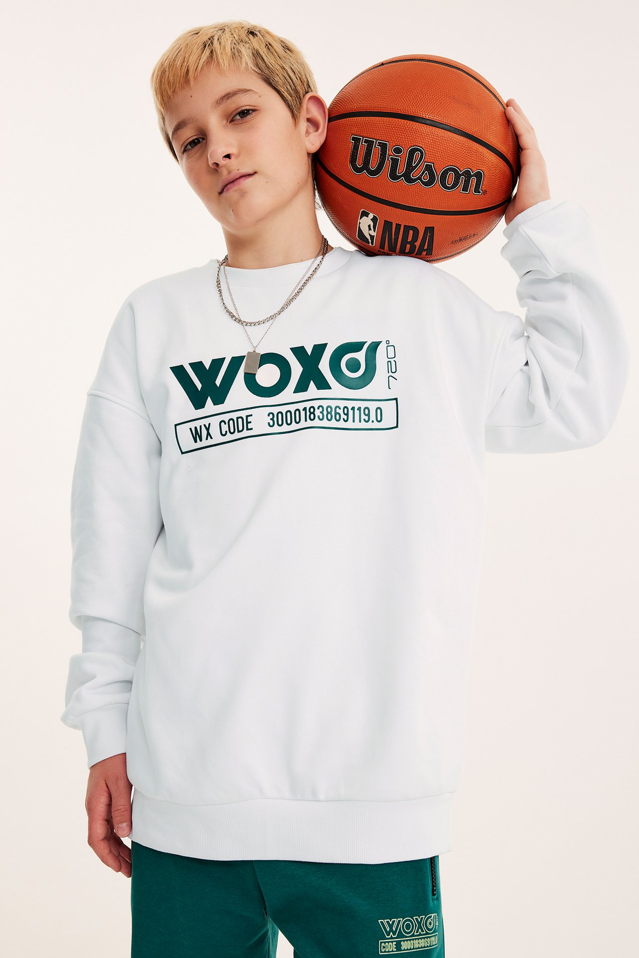 Frontvy av ett barn i en vit mönstrad sweatshirt, gröna mjukisbyxor, halsband, håller en basketboll.