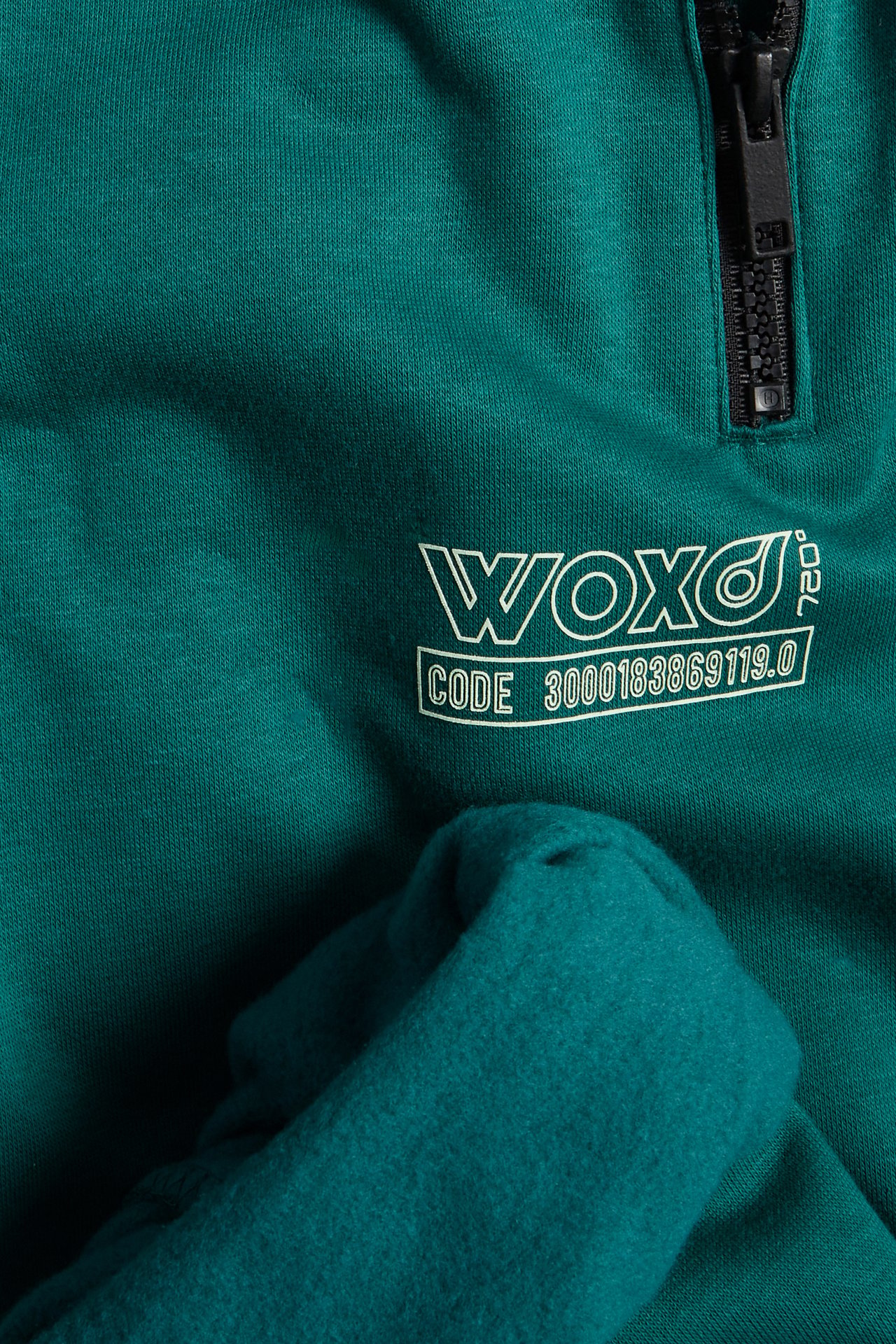 Lähikuva tummanvihreästä collegepaidasta, jossa musta vetoketju ja valkoinen "WOXO"-painatus.