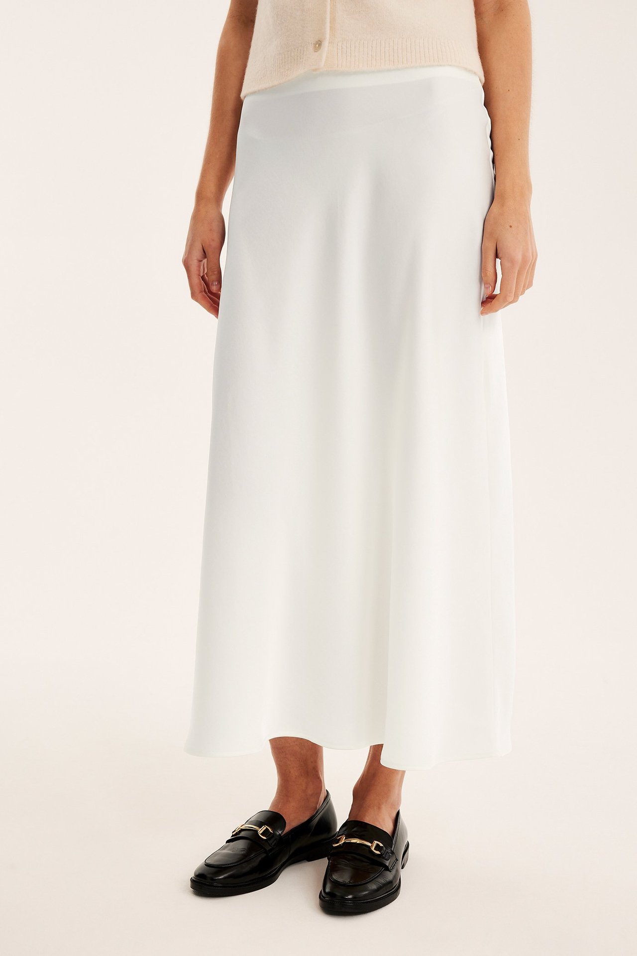 Frontbilde av en kvinne i et offwhite utsvingt satengskjørt, en lys beige cardigan og svarte loafers.
