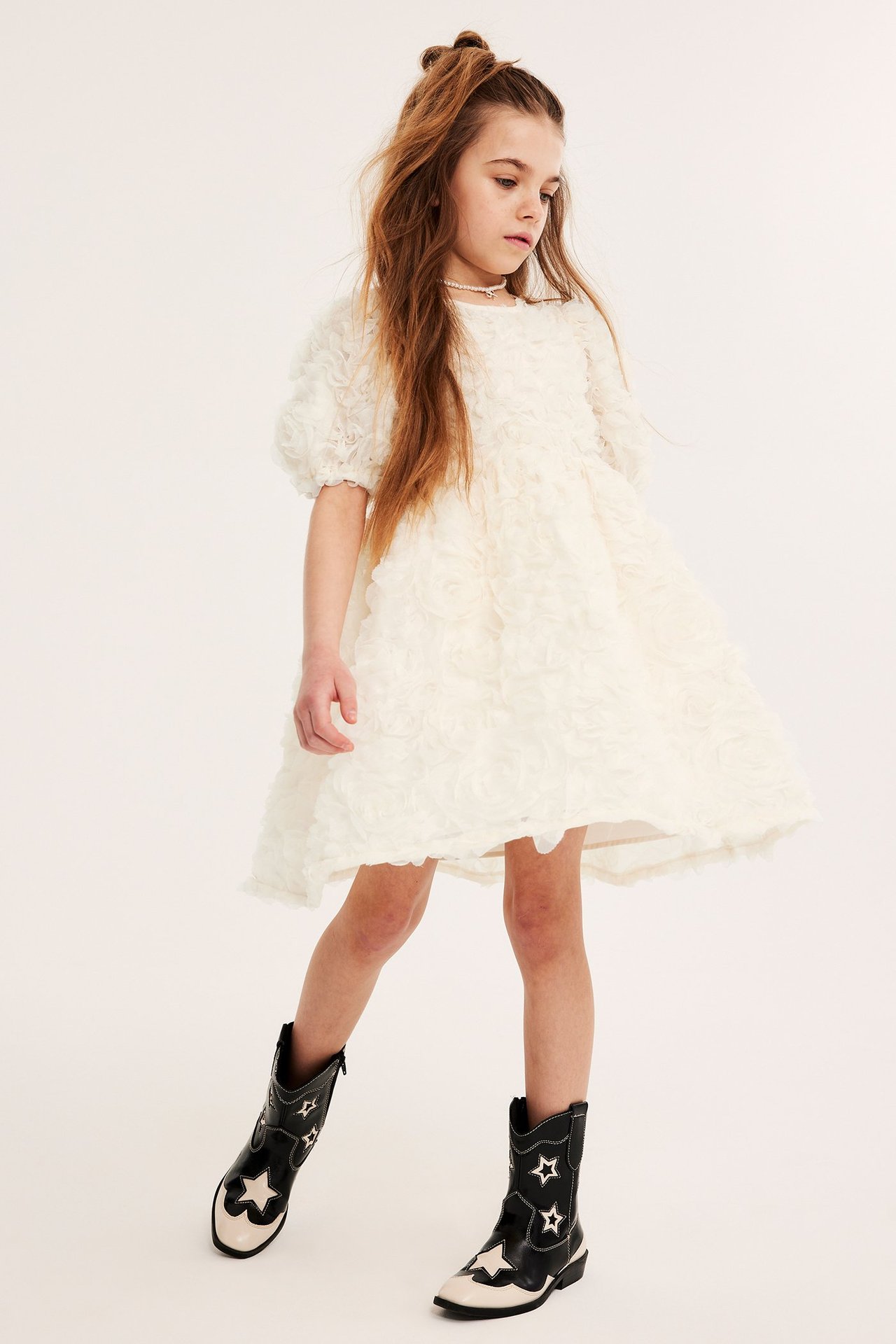 Edestä kuvattu lapsi offwhite-verkkomekossa 3D-ruusuilla, kaulakorussa ja mustavalkoisissa cowboy-saappaissa.