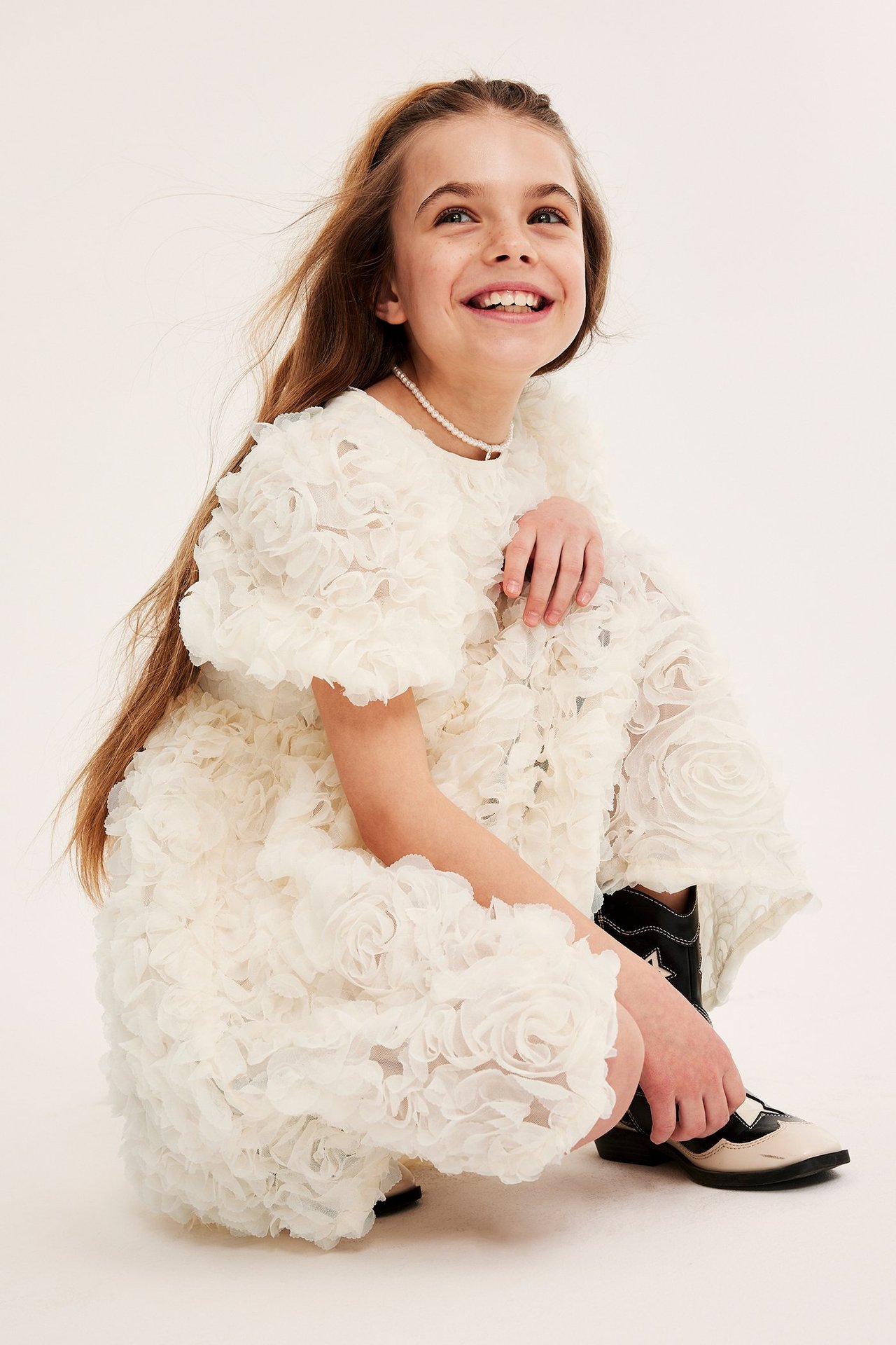 Dziecko siedzi z przodu, ubrane w sukienkę z krótkim rękawem w kwiatowy wzór. Ma długie brązowe włosy, kowbojskie buty na nogach i naszyjnik. Uśmiecha się.