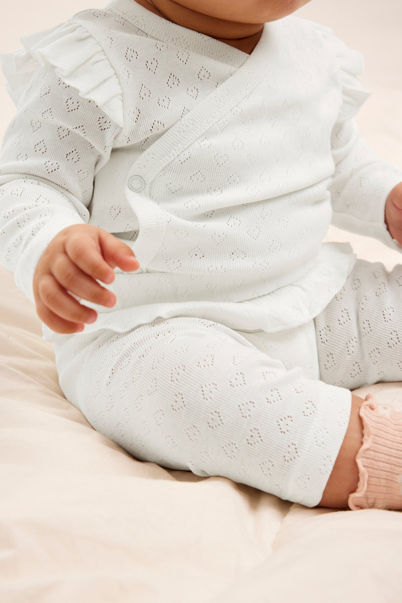 Lähikuva vauvasta: luonnonvalkoinen pointelle-body sydänkuviolla, röyhelöillä ja yhteensopivat leggingsit.