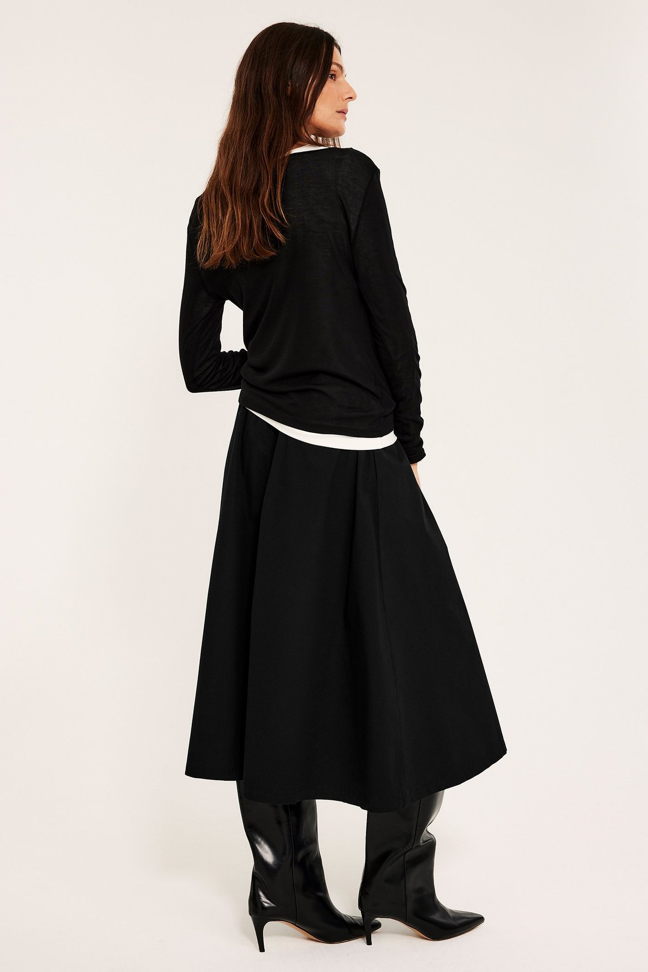 Widok z boku: kobieta w czarnej rozkloszowanej spódnicy z popeliny bawełnianej, czarnym topie z długim rękawem i czarnych butach.
