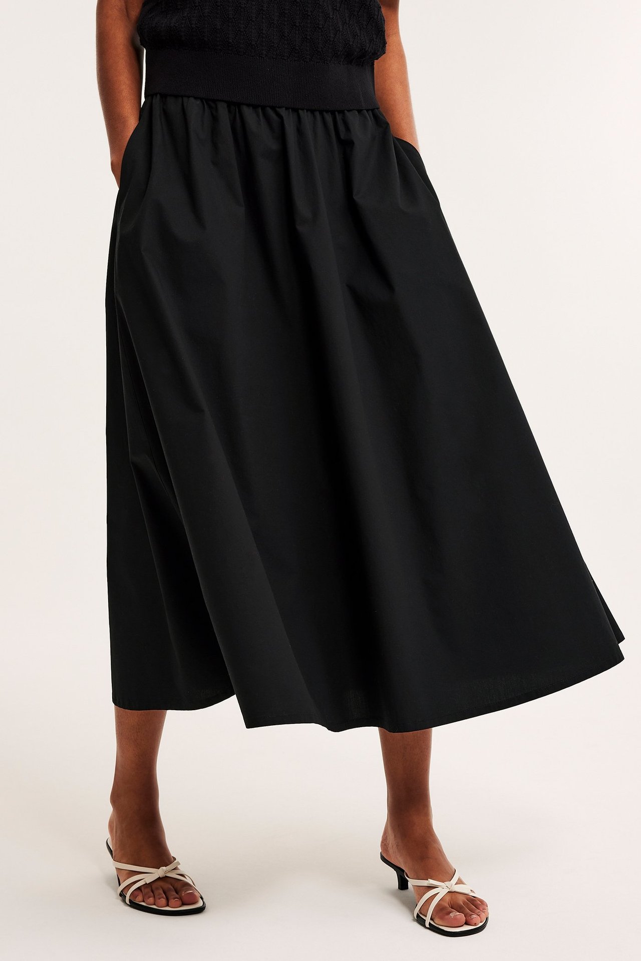 Edestäpäin, nainen mustassa levenevässä popliinihameessa, mustassa topissa ja valkoisissa korkokengissä.