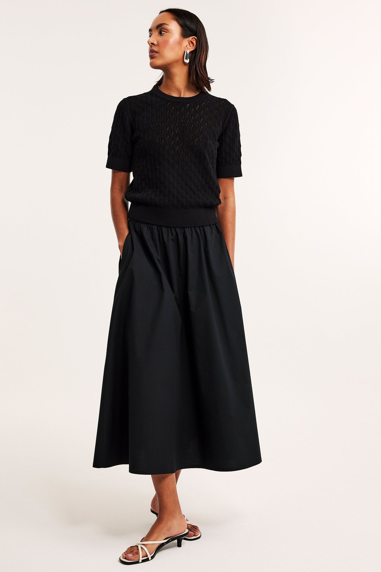 Widok z przodu: Kobieta w czarnej rozkloszowanej spódnicy poplinowej z gumką, czarnym wzorzystym topie, białych sandałach.