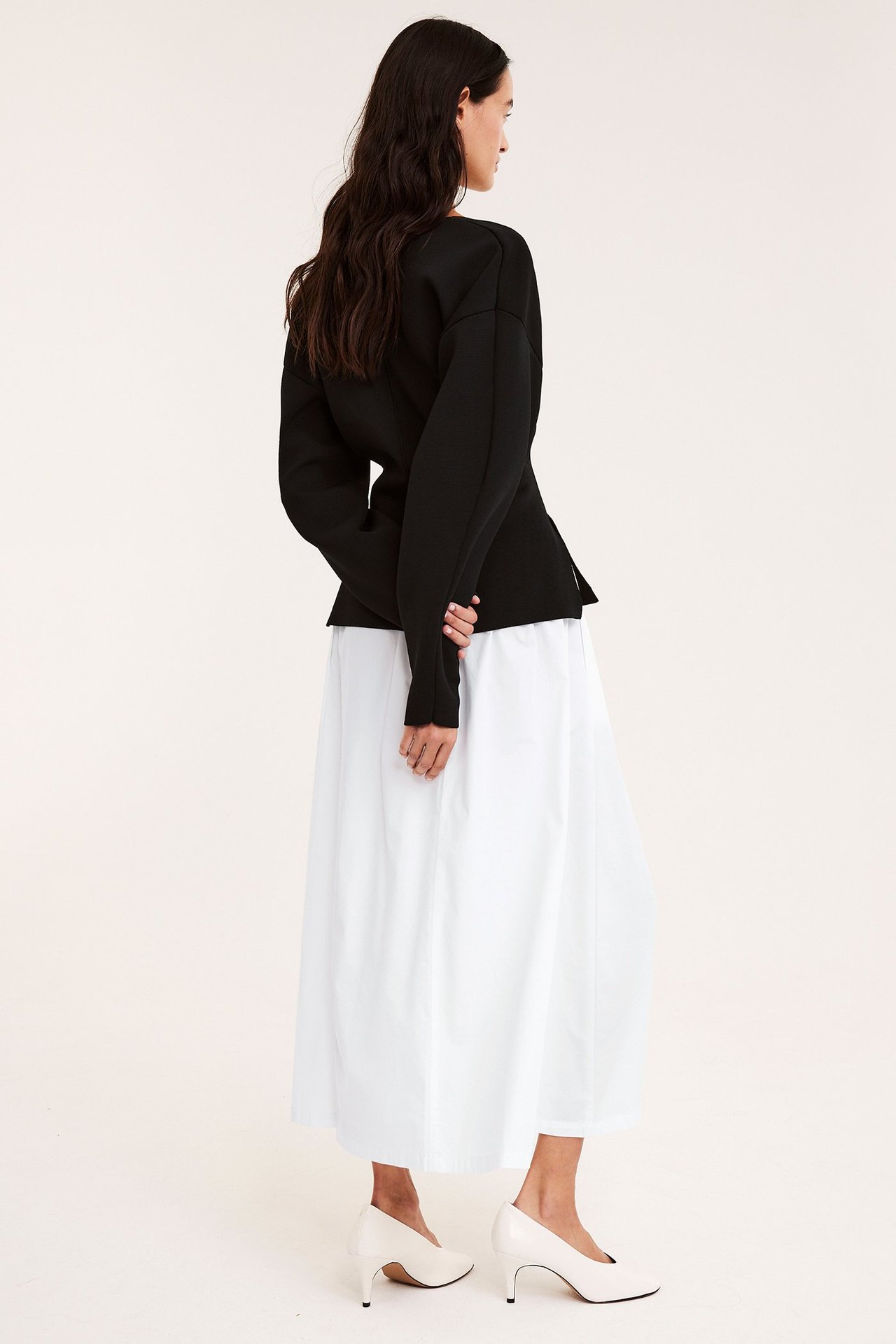 Sidovy av kvinna i vit utställd kjol i bomullspoplin, svart långärmad topp och ljusbeige pumps.