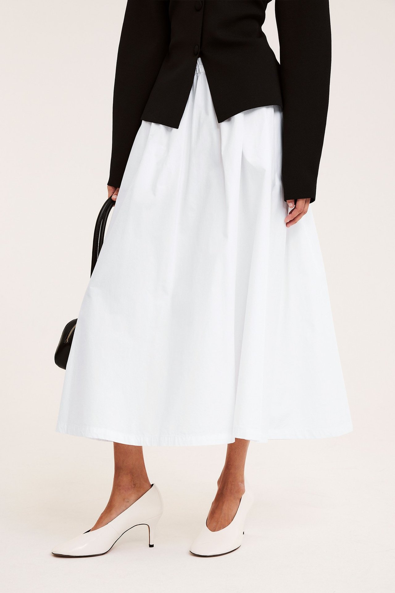 Forfra: Kvinne i hvitt bomullspoplin skjørt, svart jakke, hvite pumps, med svart veske.