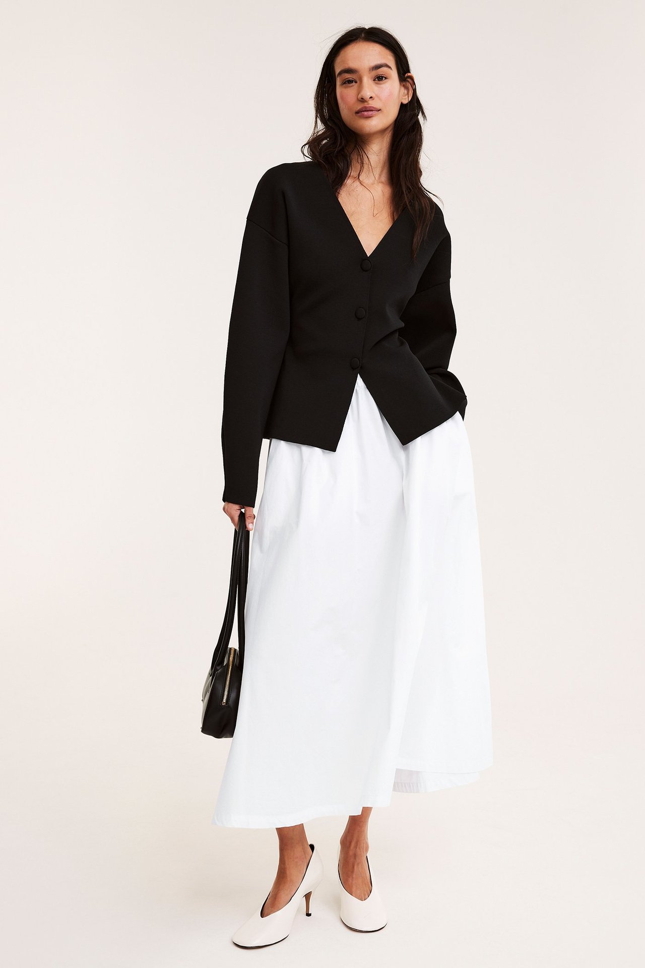 Framsida: Kvinna i vit utställd bomullspoplinkjol, svart V-ringad topp, svart handväska och vita klackar.