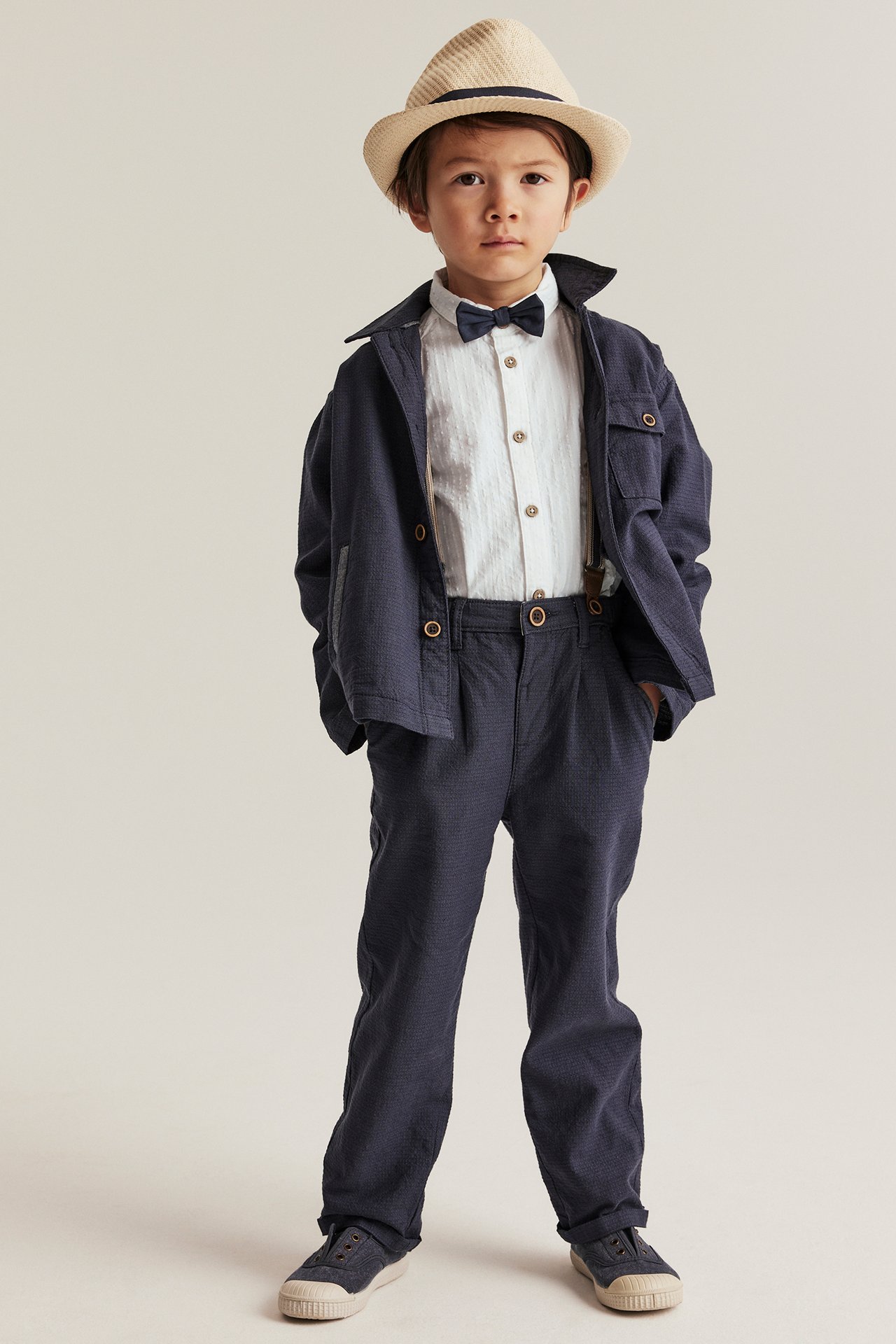 Frontbilde av et barn: offwhite vevd skjorte, mørkeblå sløyfe, jakke, bukser og en beige hatt.
