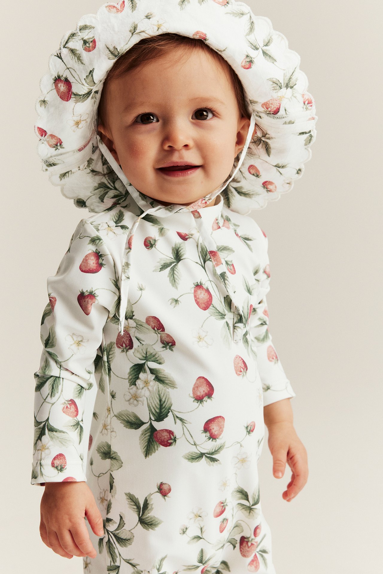 Edestä, vauva yllään luonnonvalkoinen mansikkakuvioinen pitkähihainen UV-asu ja yhteensopiva aurinkohattu.