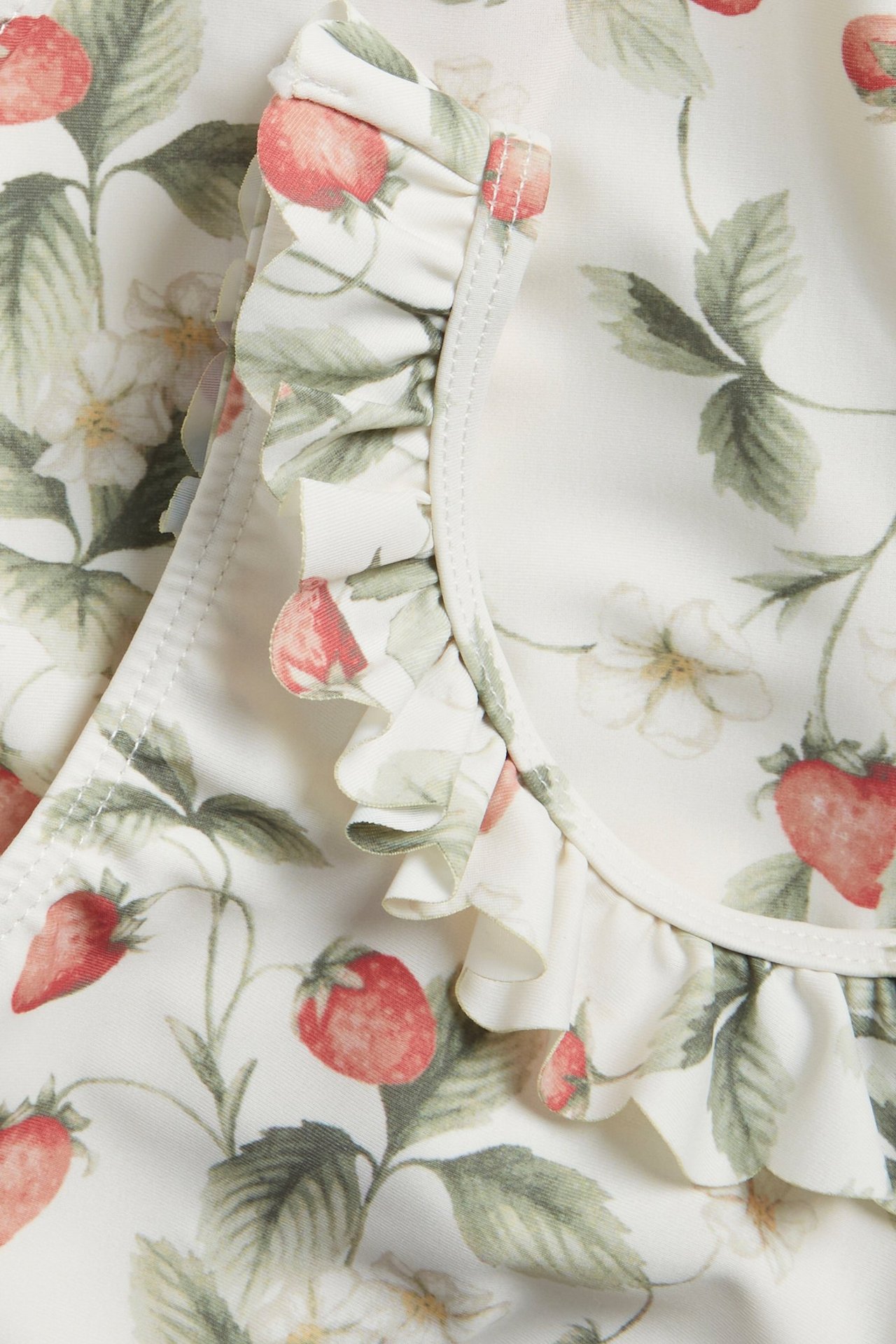 Lähikuva luonnonvalkoisesta lasten uimapuvusta, jossa mansikkakuvio ja röyhelöt pääntiessä.