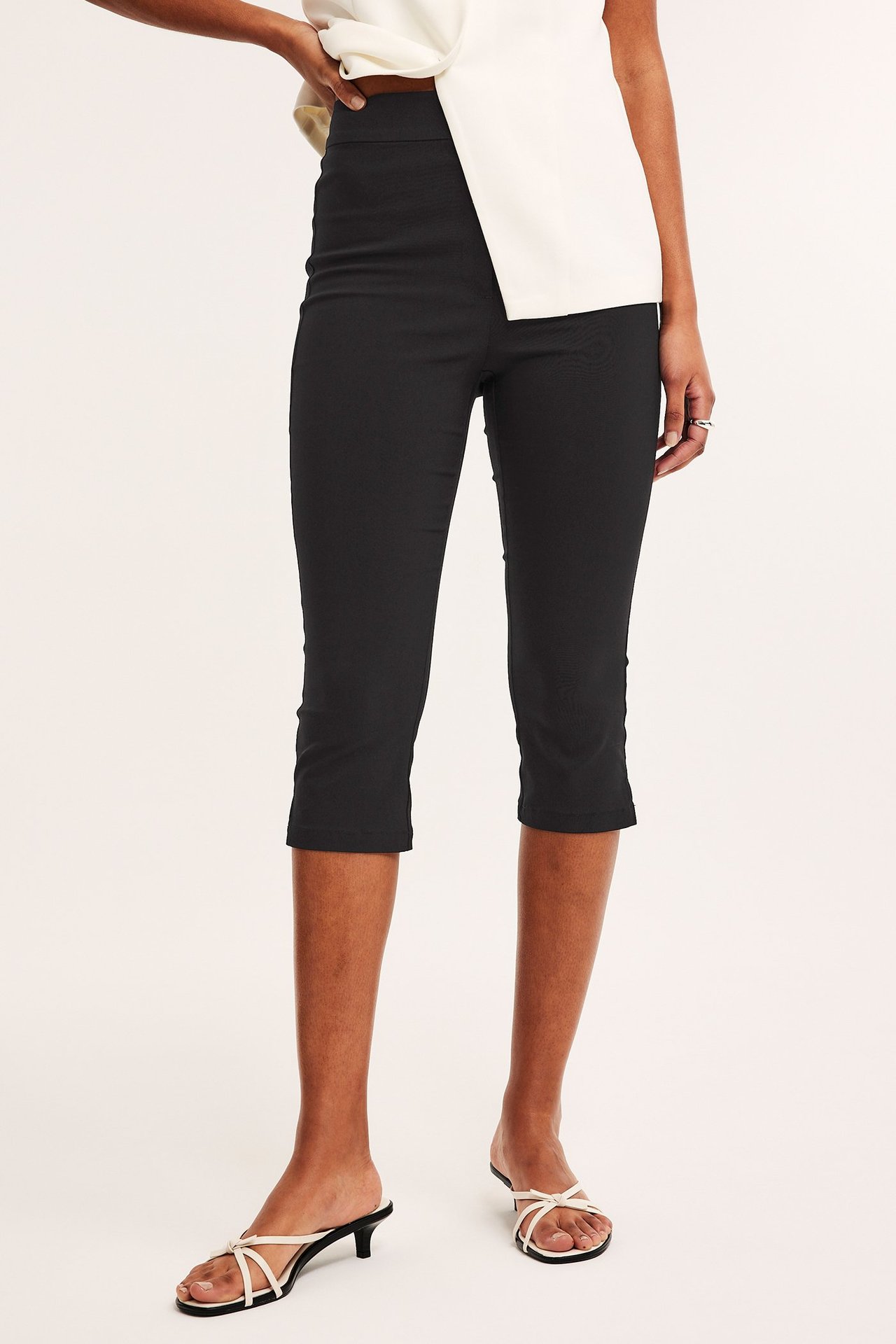 Edestä kuvattu nainen mustissa bengaliini-caprihousuissa, valkoisessa epäsymmetrisessä topissa ja valkoisissa sandaaleissa.